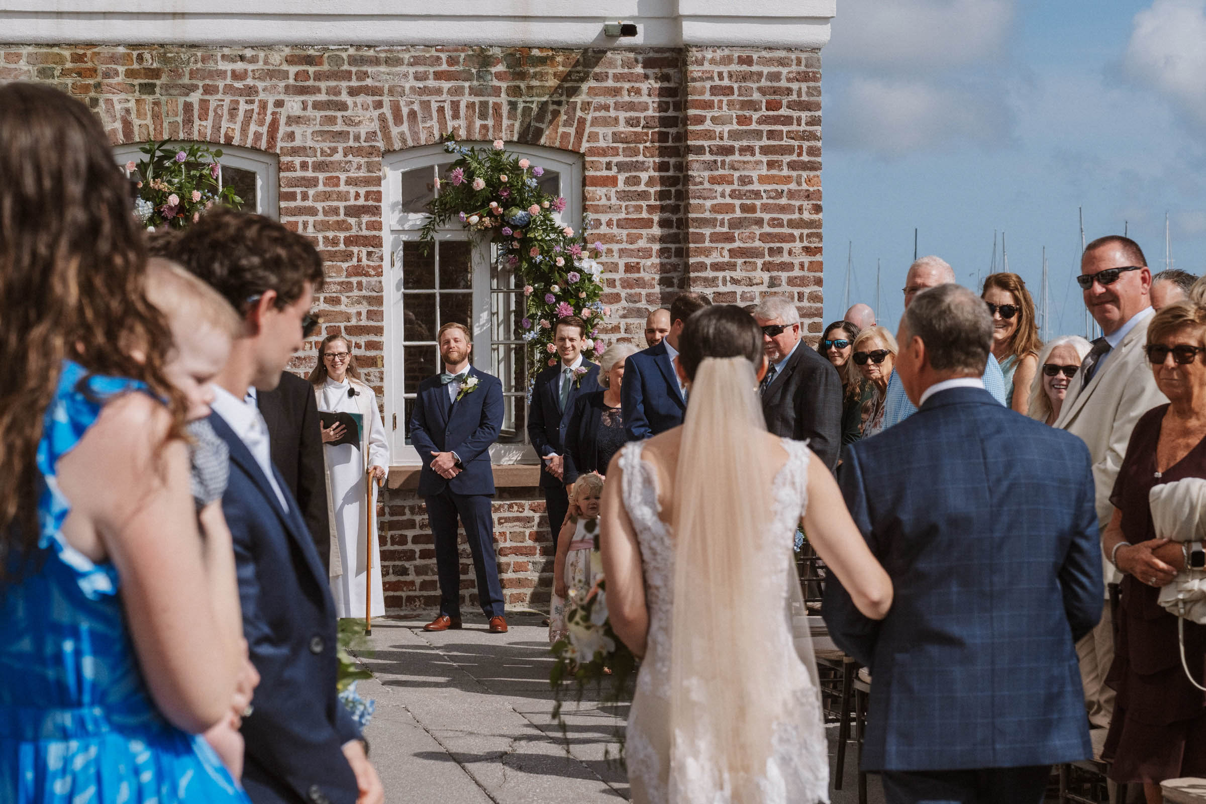 Historic Rice Mill Courtyard Ceremony Bride walks toward the ceremony with a family member beside the brick Historic Rice Mill and marina.