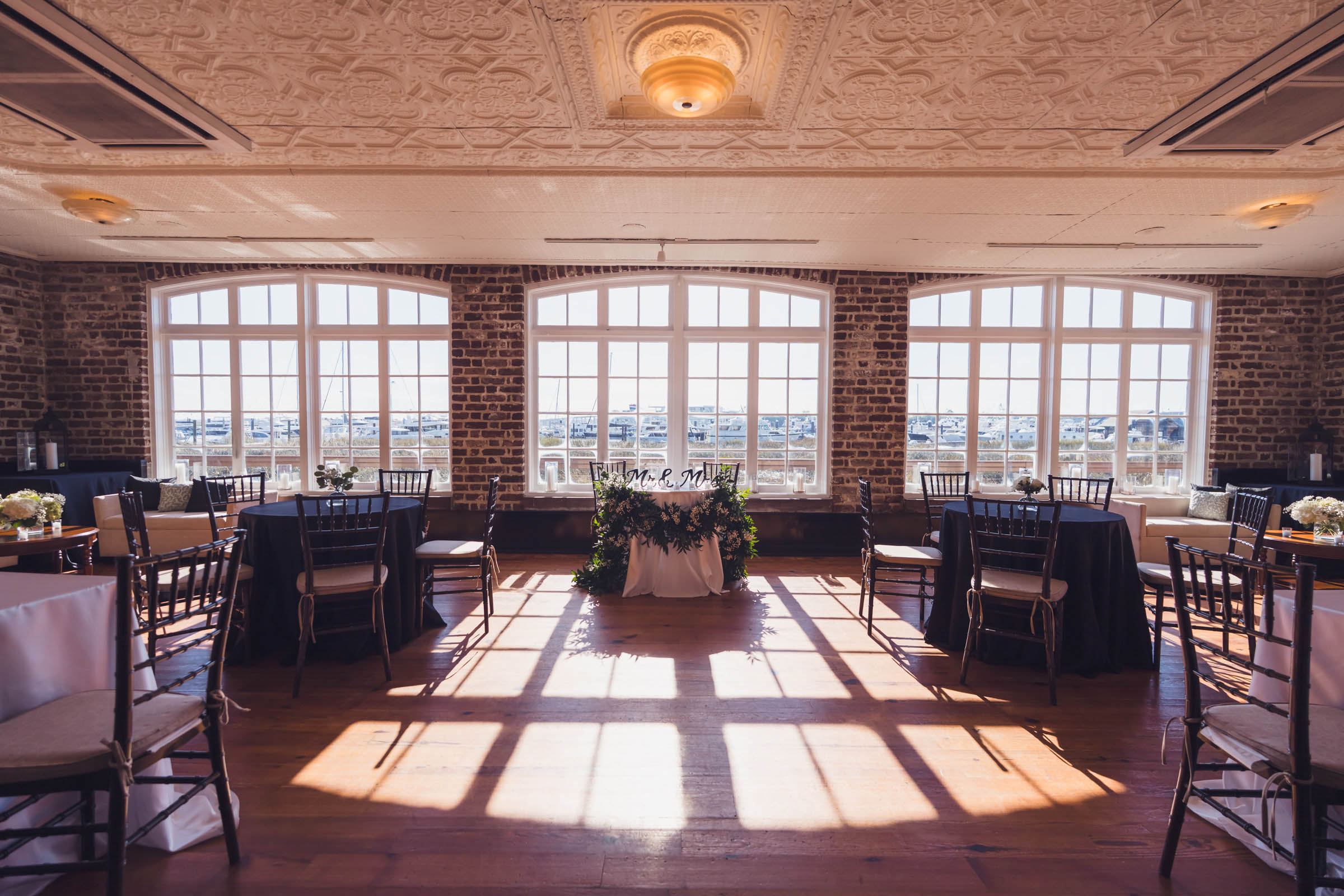 Historic Rice Mill Marina Room Sunlight streams through the large waterfront windows of the Historic Rice Mill reception room with tables set for a wedding.