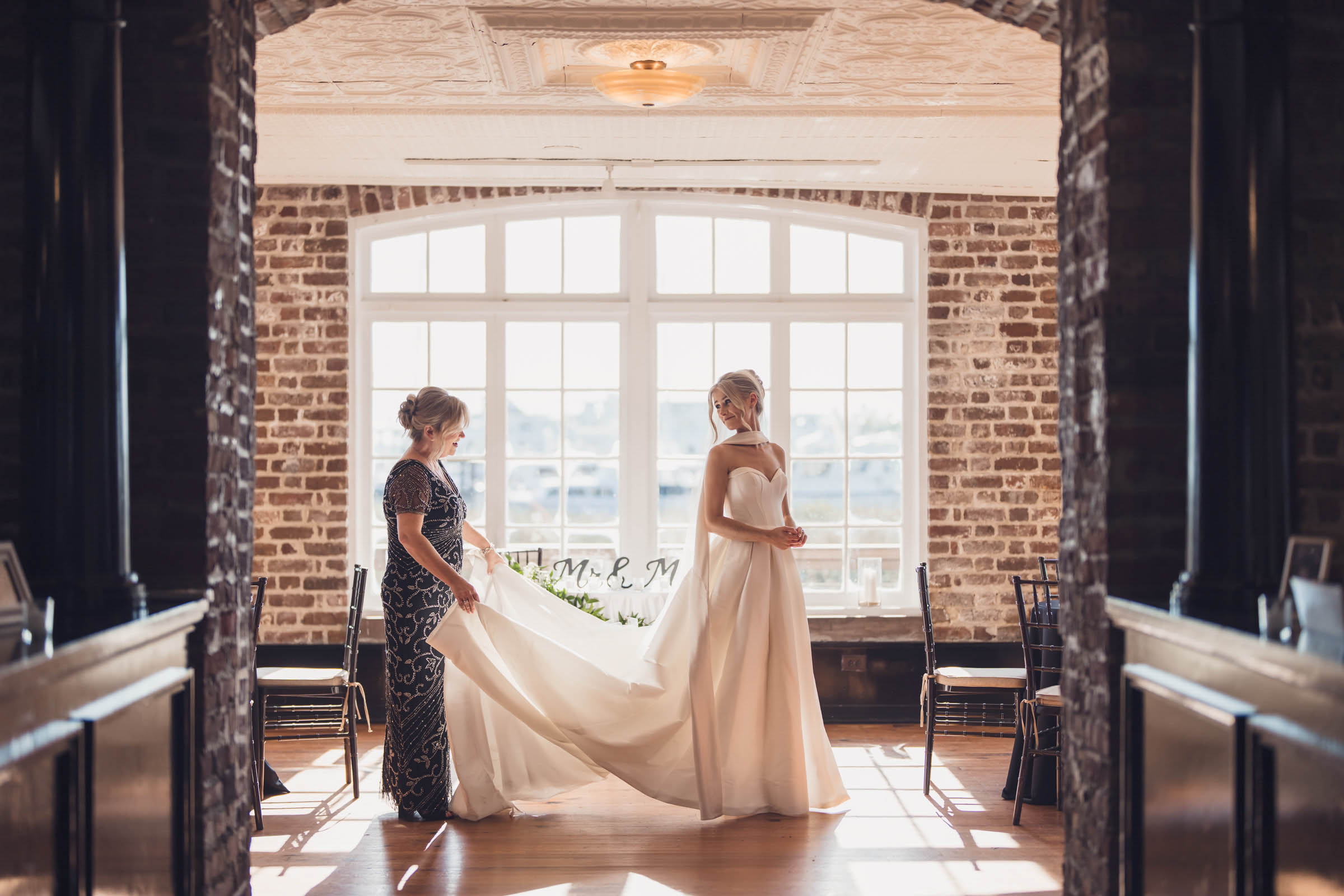 Bride Getting Ready at the Historic Rice Mill Bride stands in her wedding gown while a family member arranges the train beside the large windows of the Historic Rice Mill.