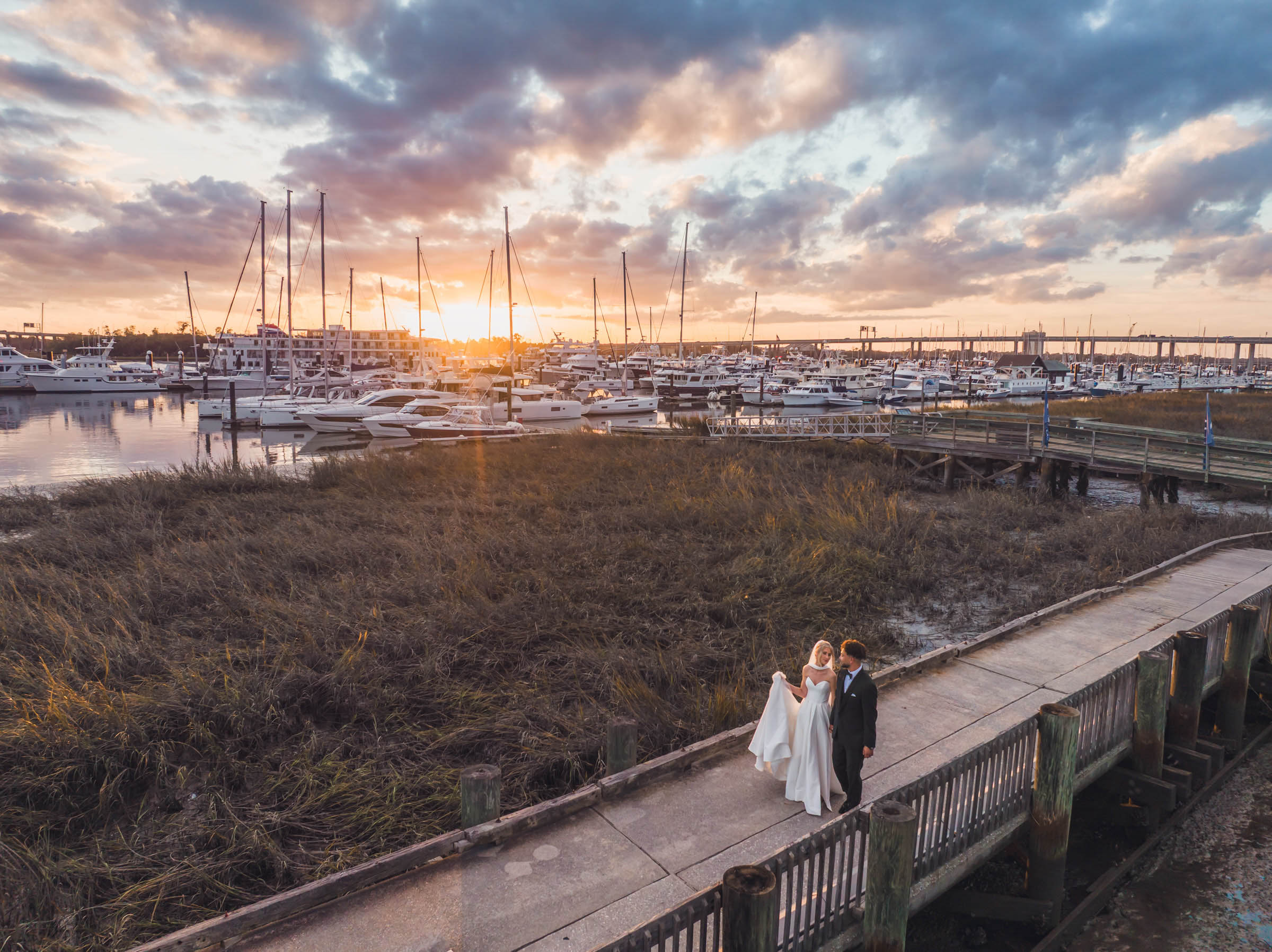 Historic Rice Mill Waterfront Sunset Portrait Newlyweds walk along the waterfront beside the marina beneath a colorful Charleston sunset near the Historic Rice Mill.