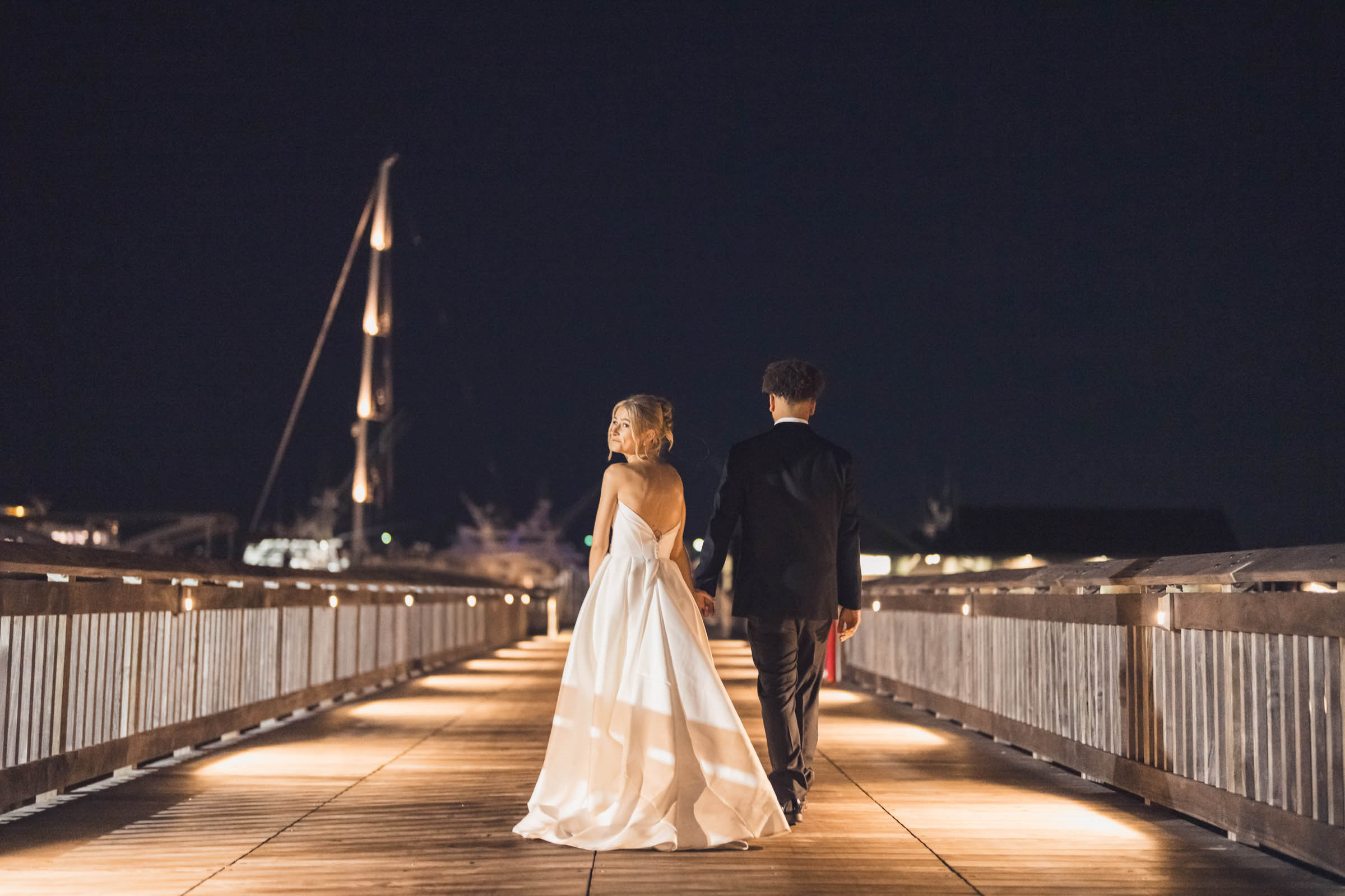 Historic Rice Mill Night Waterfront Portrait Newlyweds walk hand in hand along the illuminated waterfront boardwalk after their Historic Rice Mill wedding.