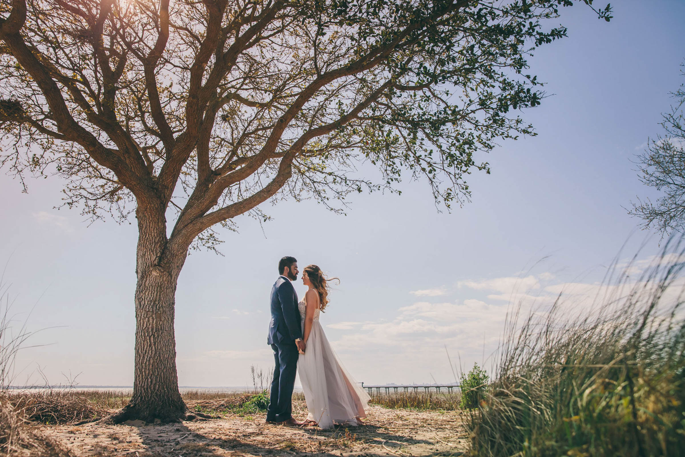 Bride and groom holding hands beneath a windswept tree by the marsh, with coastal grasses and a distant pier—romantic Charleston outdoor wedding portrait.