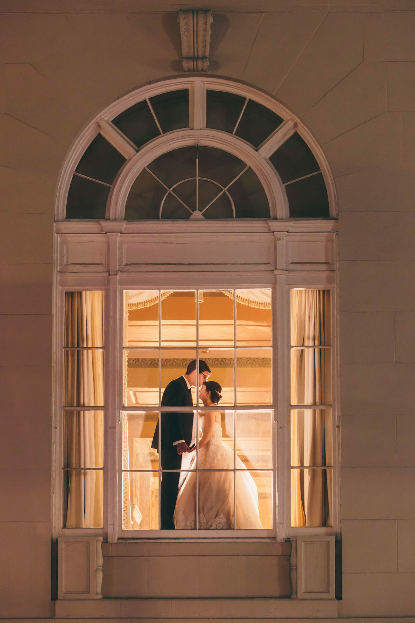 Bride and groom standing close together framed by a large arched window glowing with warm indoor light—elegant nighttime Charleston wedding portrait.