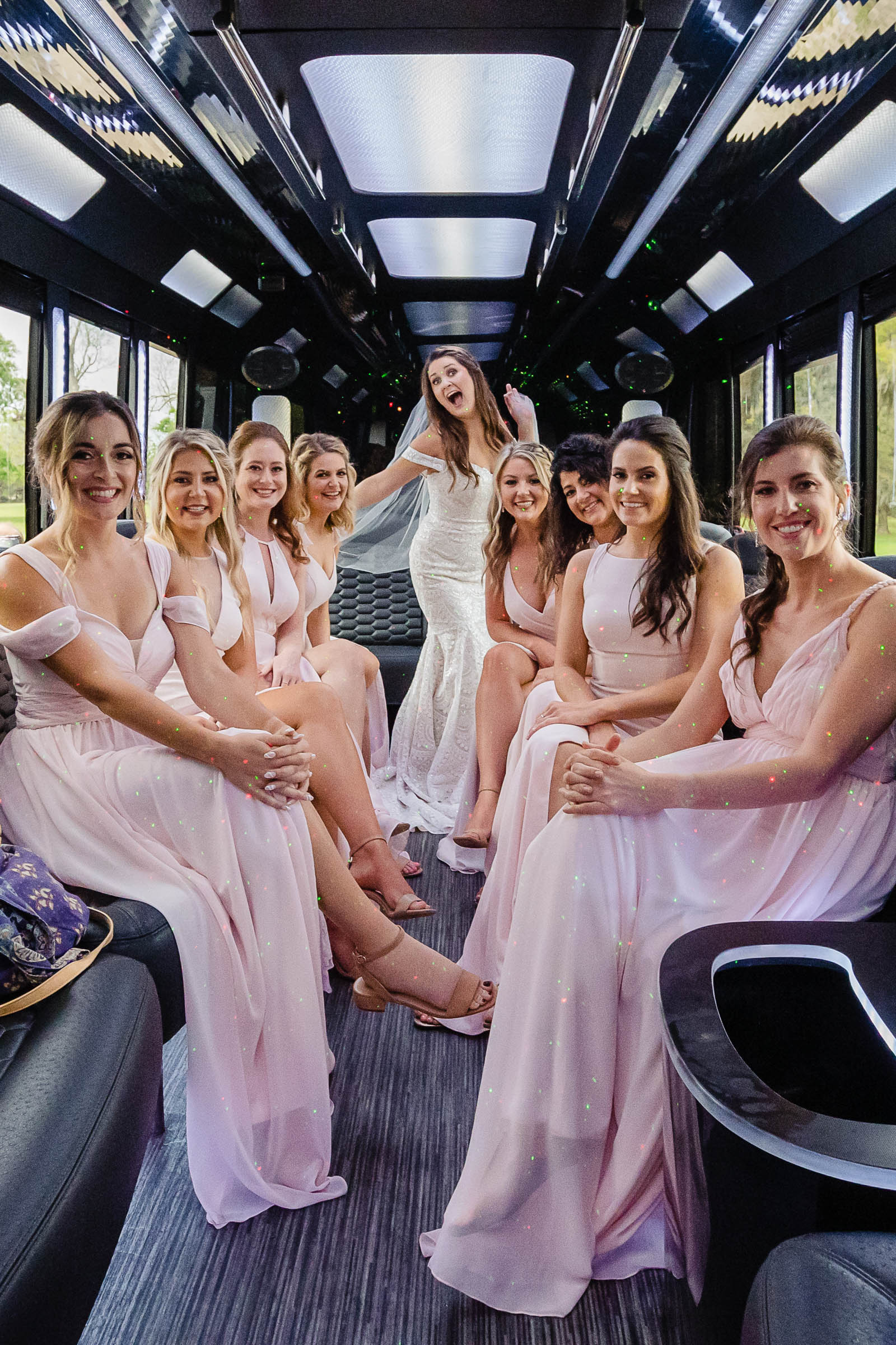 Bride and bridesmaids in blush dresses celebrating inside a party bus with colorful lights—fun candid wedding day moment with bridal party.