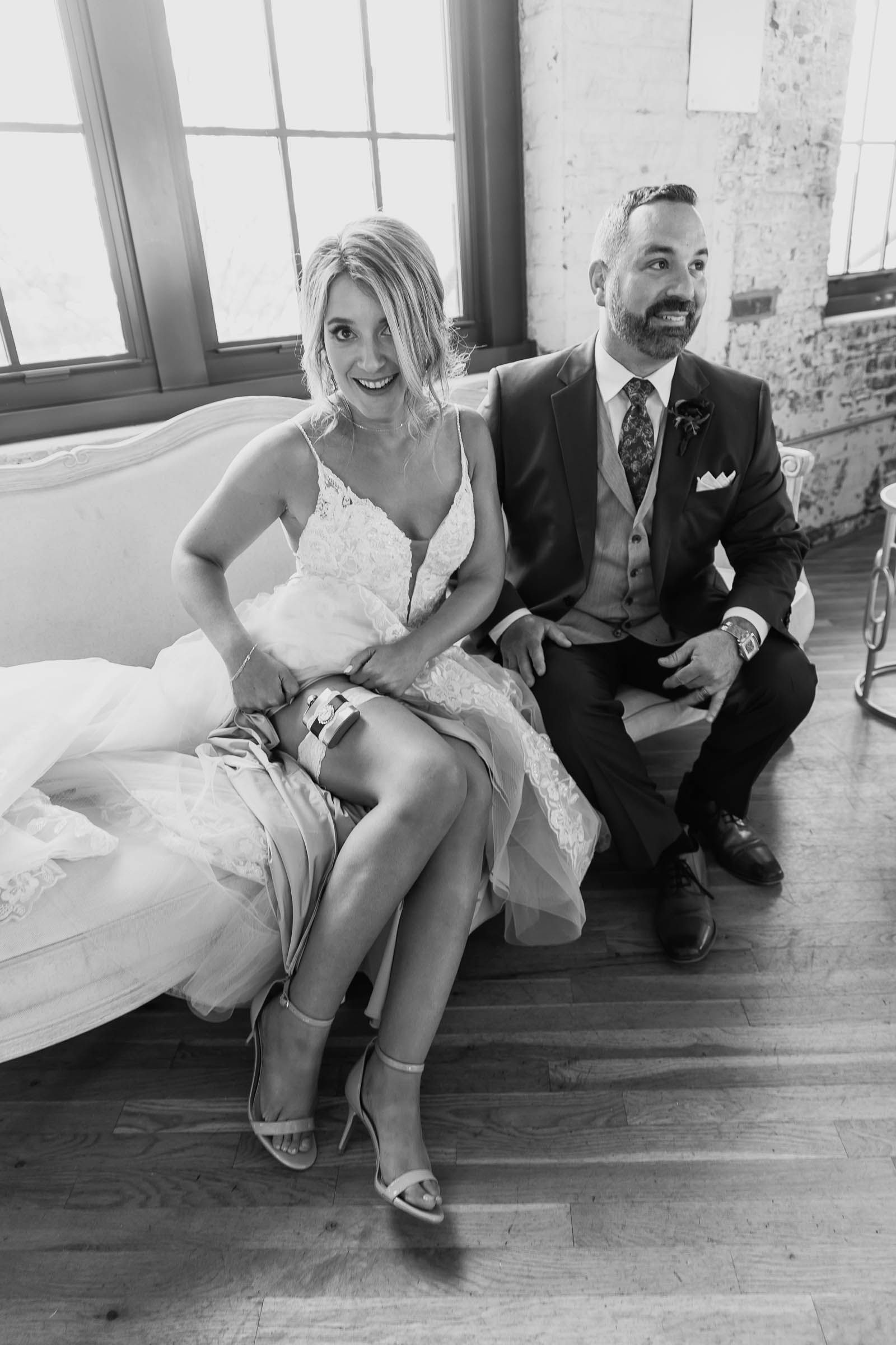 Bride laughing while adjusting her garter on a vintage sofa beside the groom in a sunlit loft with large windows—candid black and white wedding moment.
