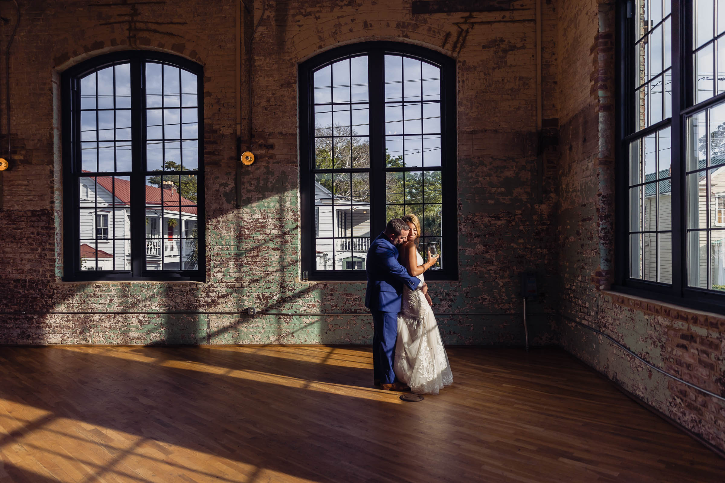 Bride and groom embracing in a sunlit industrial loft with large arched windows and textured brick walls—romantic Charleston wedding portrait.