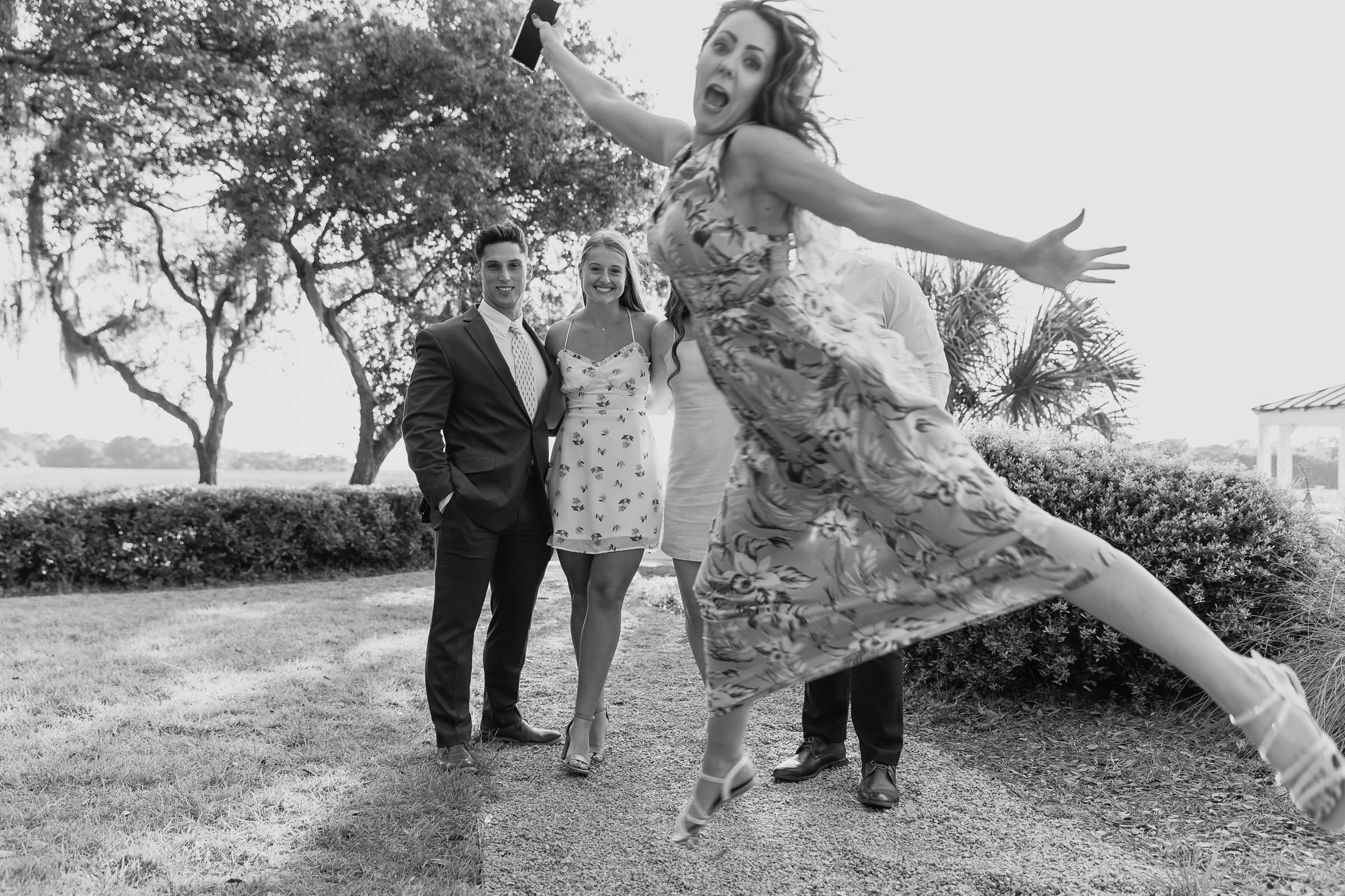 Wedding guest jumping into the frame mid-air in front of a smiling couple outdoors with oak trees and Spanish moss—playful candid Charleston wedding photo.