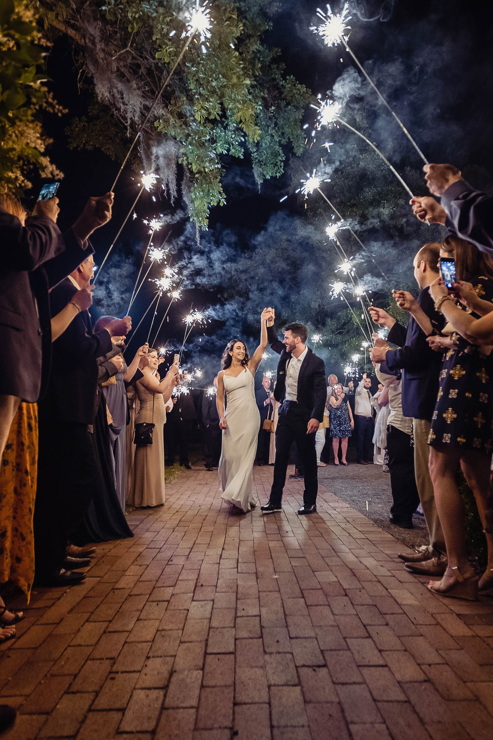 Newlyweds walking through a sparkler exit at night surrounded by cheering guests—joyful Charleston wedding send-off on a brick pathway.