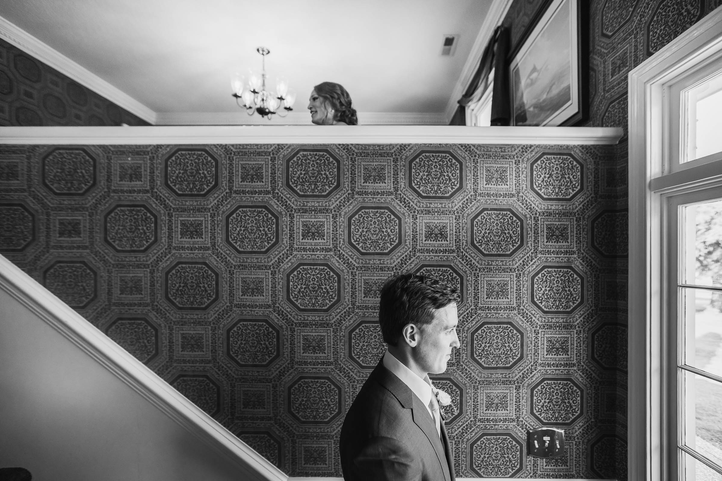 Groom standing by a window in a patterned stairwell while the bride appears above on the landing—artistic black and white wedding moment with layered composition.