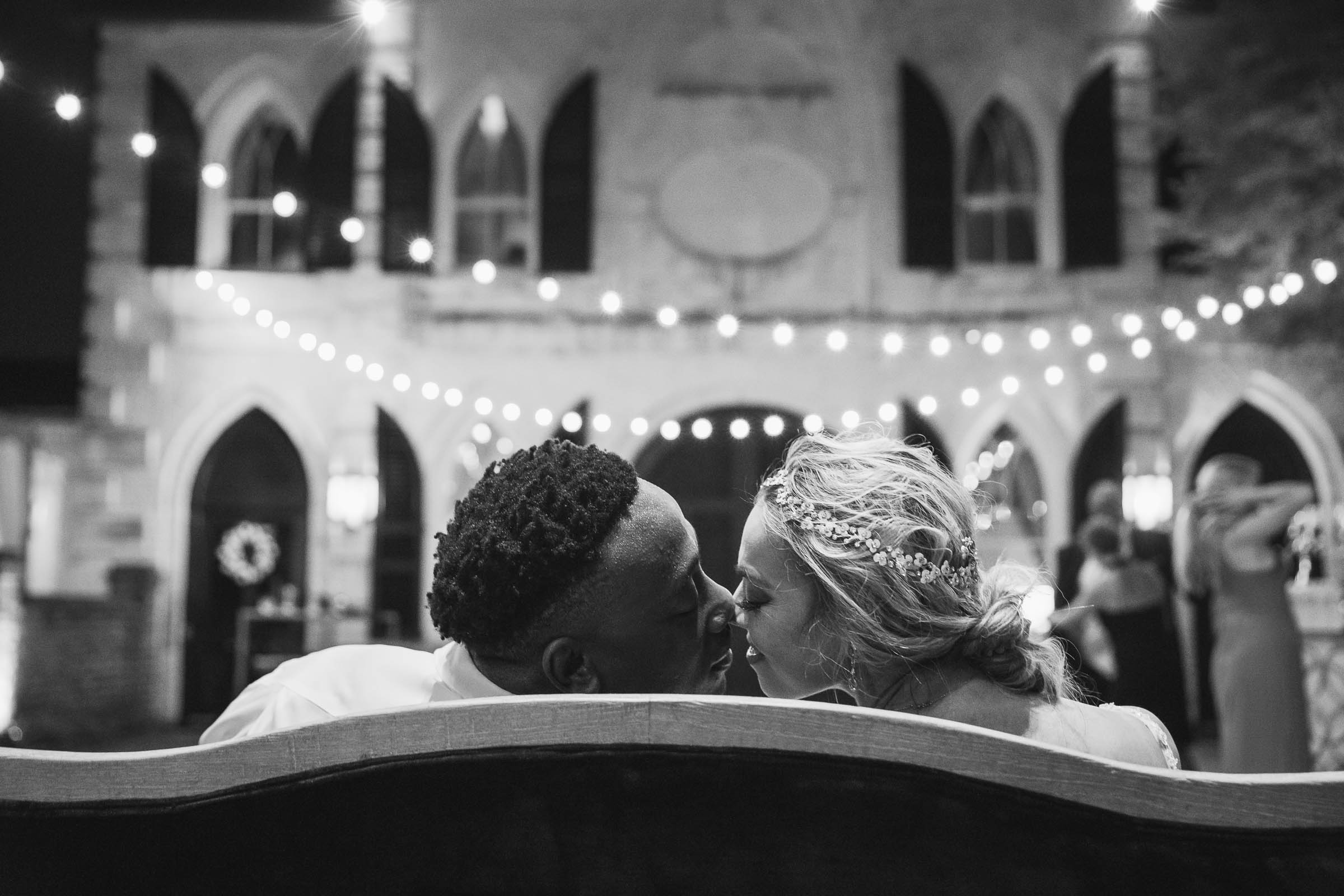 Bride and groom leaning in for a kiss on a bench beneath string lights in a historic courtyard—romantic black and white Charleston wedding photo.