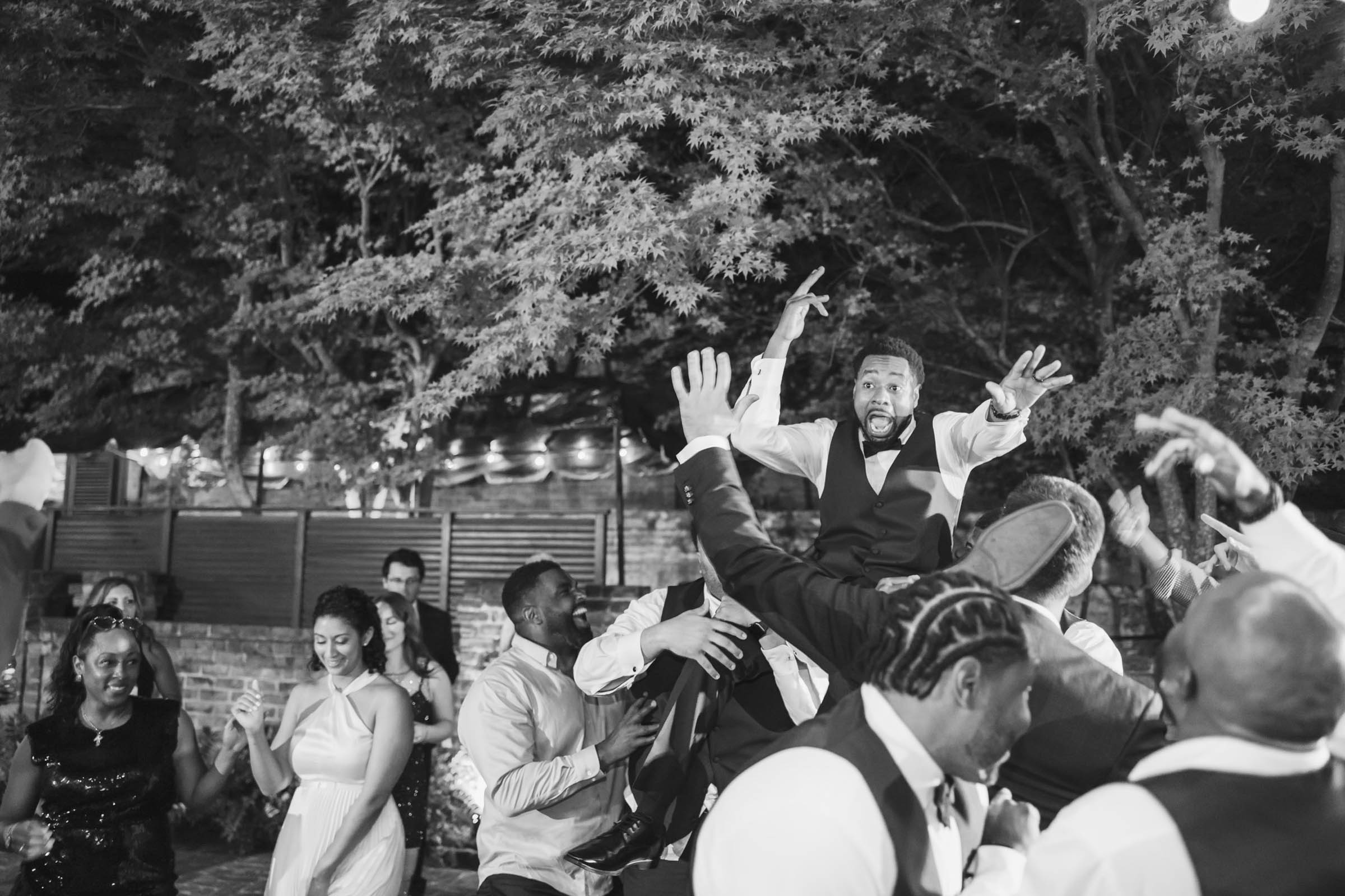 Groom being lifted into the air by friends on the dance floor during an outdoor reception—energetic black and white wedding celebration.