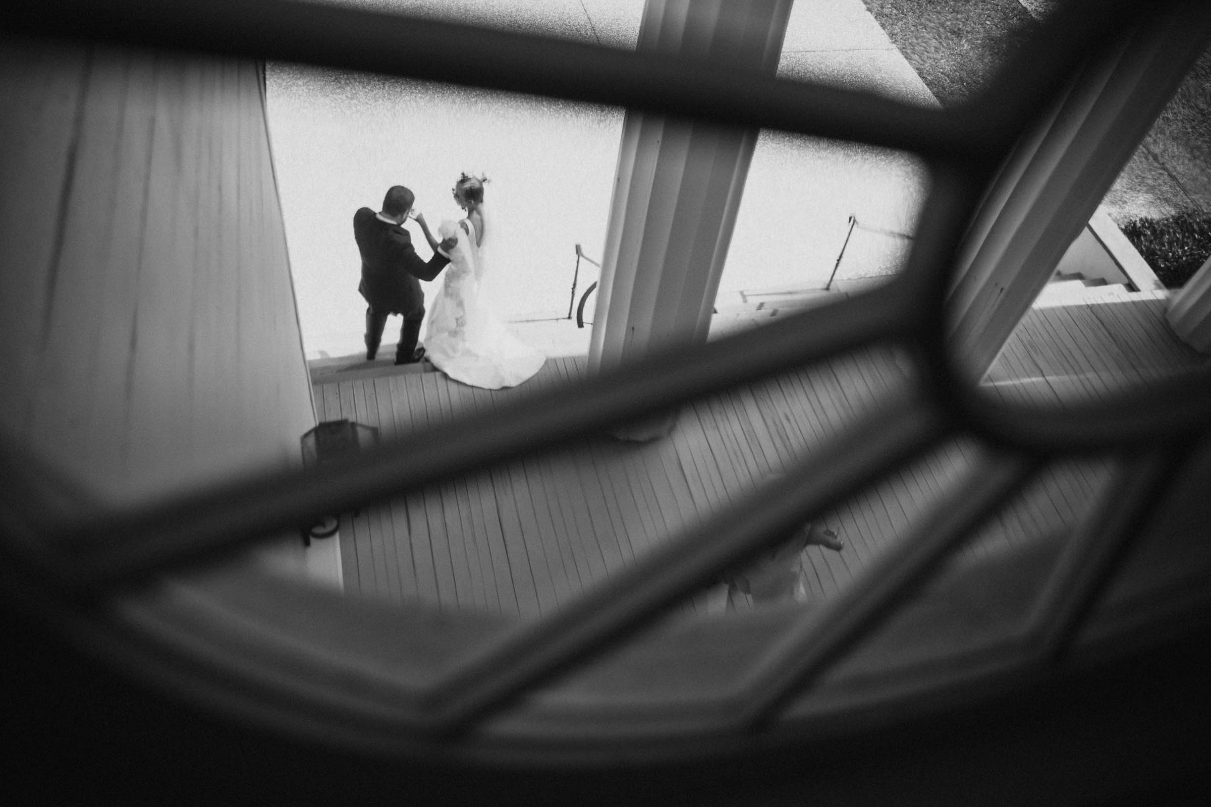 Bride and groom descending exterior steps, photographed from inside through a second-floor window—framed by window lines in a black and white composition.