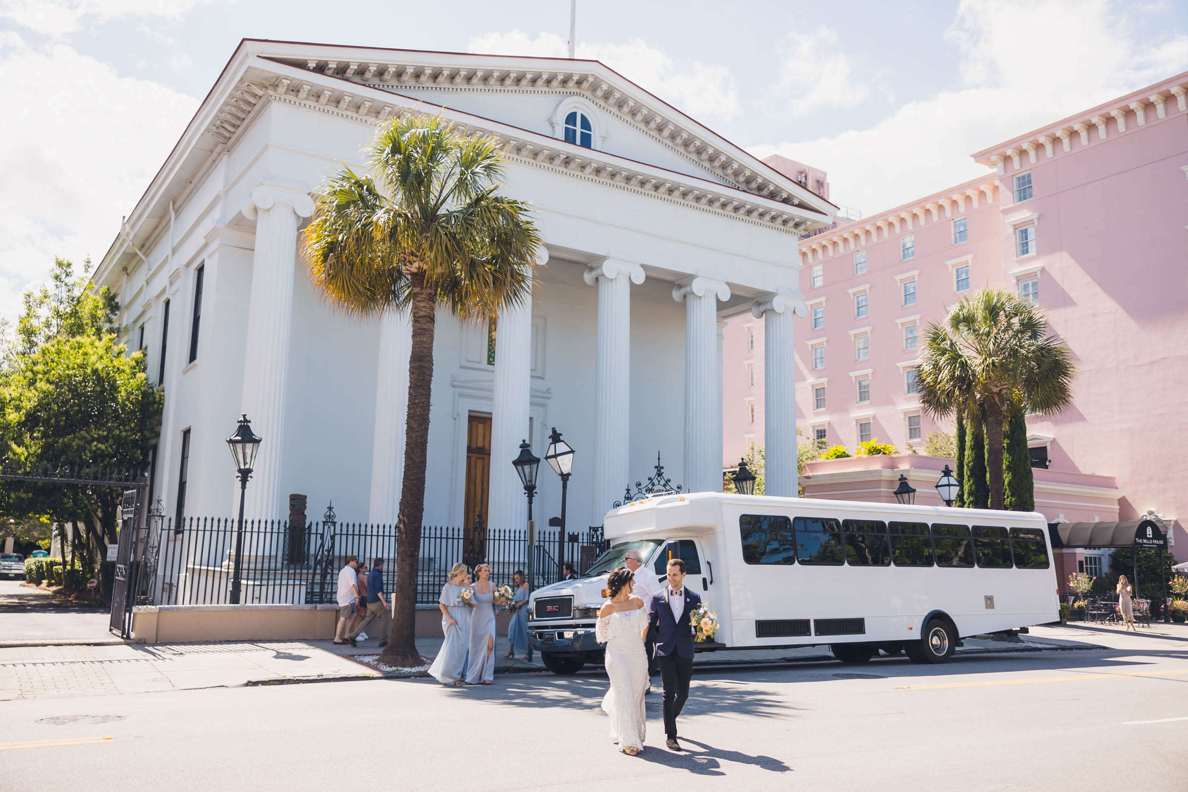 Bride and groom walking across the street in front of a historic Charleston Hibernian Society Hall with a white party bus and wedding party nearby—sunlit downtown wedding scene.