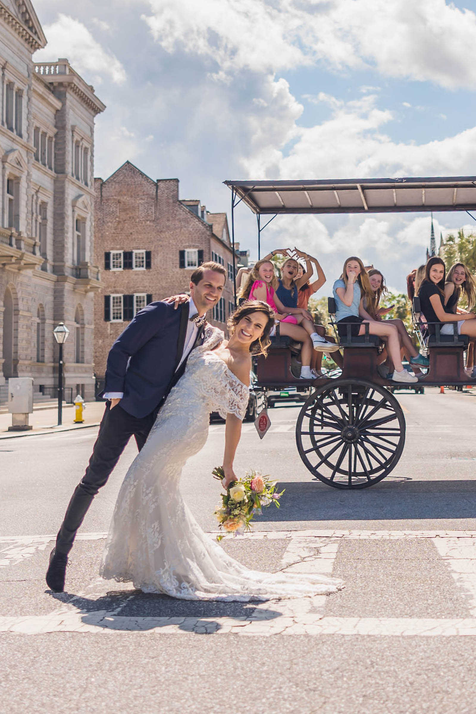 Bride and groom posing playfully in the street with a horse-drawn carriage. A teenager in the background flashes an expert heart hands as she photobombs.