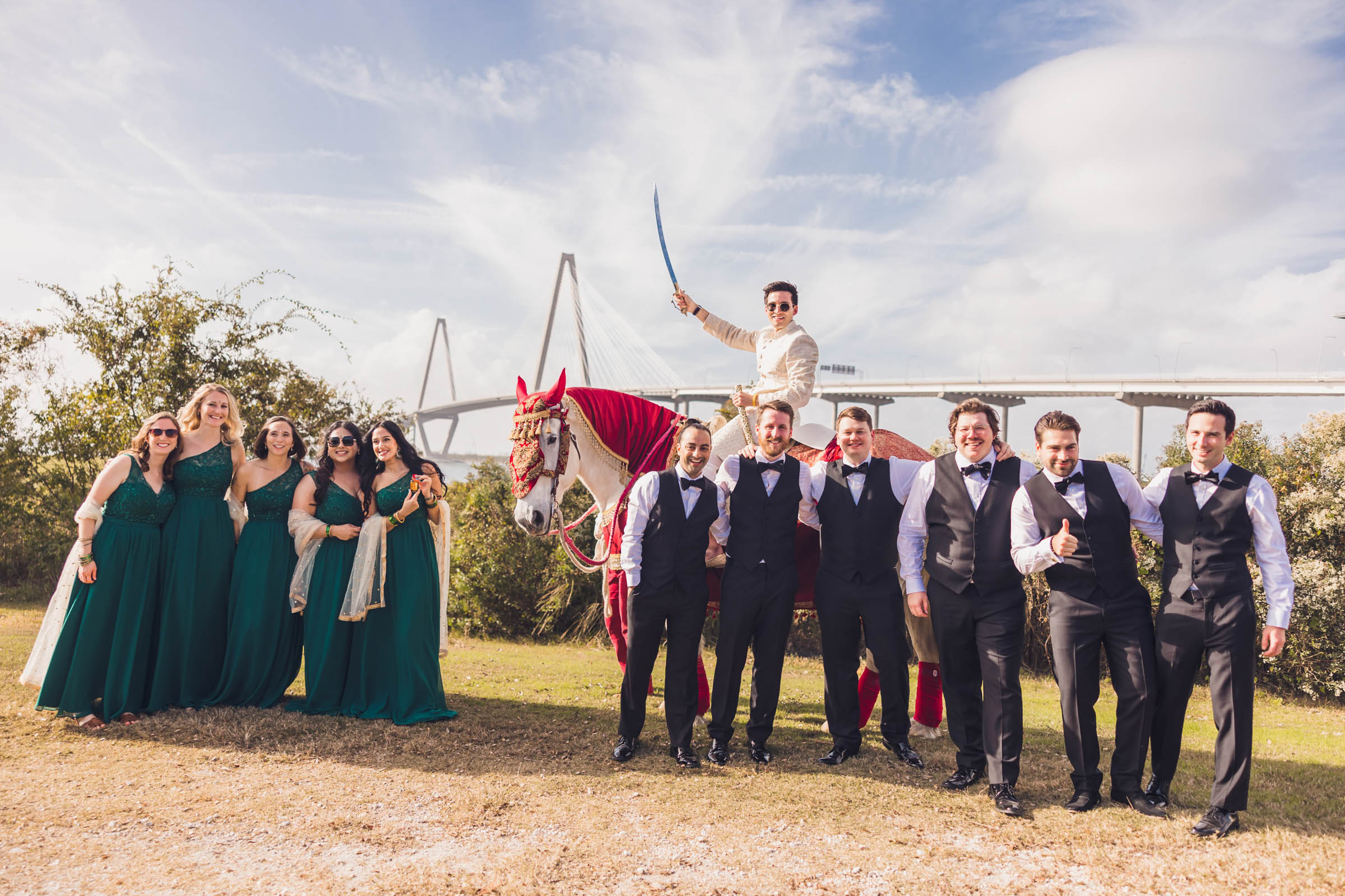 Groom riding a decorated horse holding a ceremonial sword with wedding party posing nearby and the Ravenel Bridge in the background—bold Charleston wedding portrait.