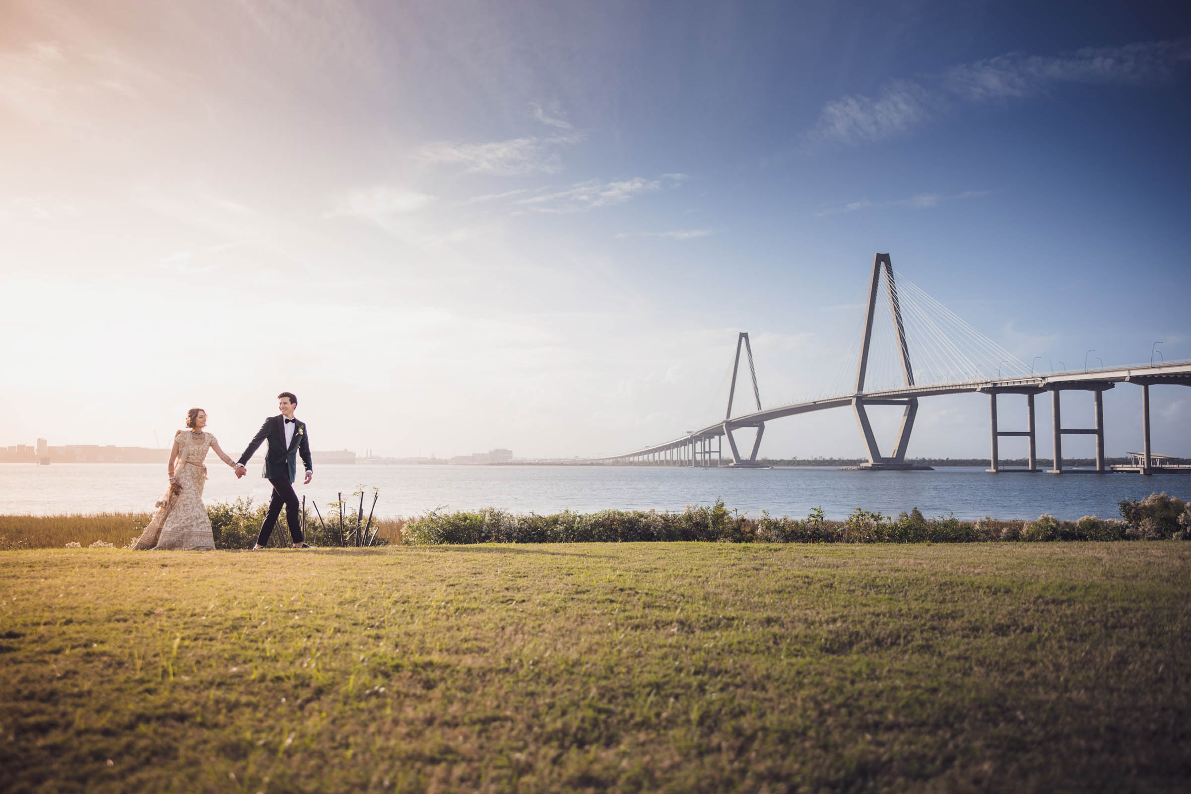 Bride and groom walking hand in hand along the waterfront with the Ravenel Bridge stretching across the background—wide, sunlit Charleston landscape.
