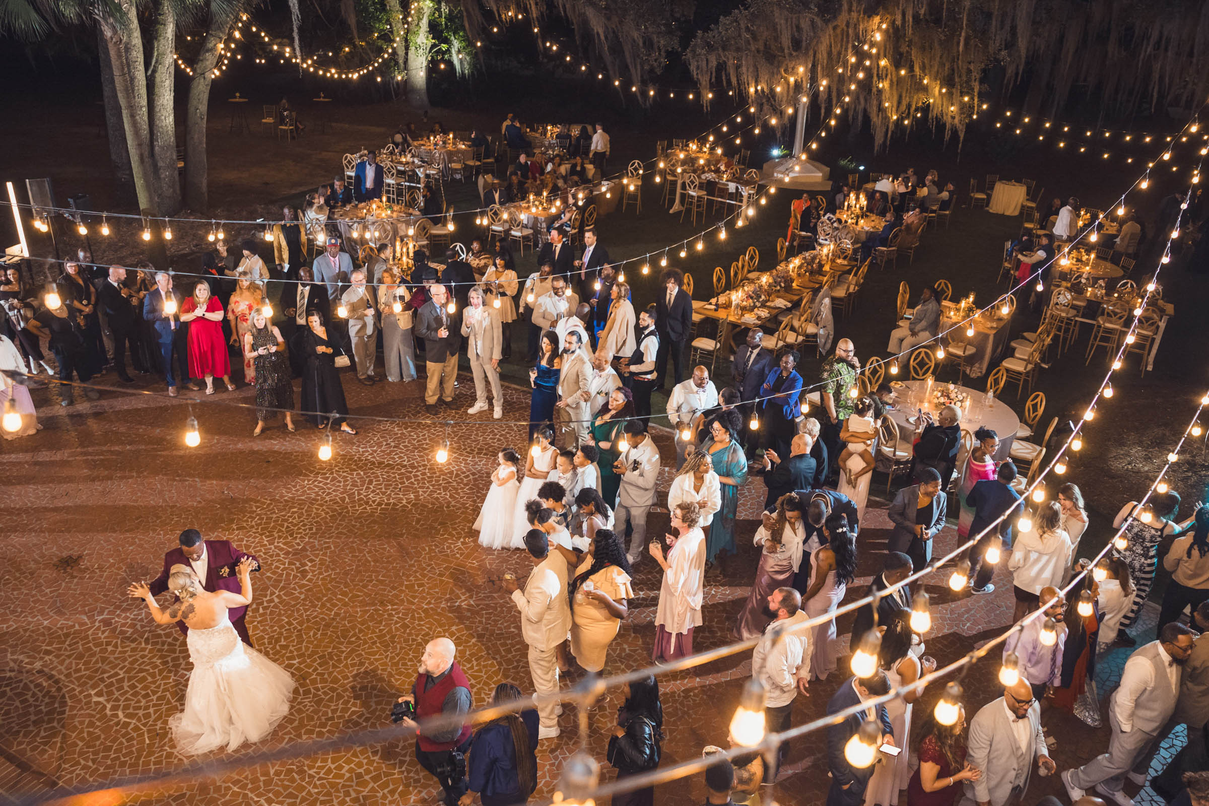 Overhead view of a lively outdoor wedding reception with guests dancing under string lights and oak trees—Charleston evening celebration.