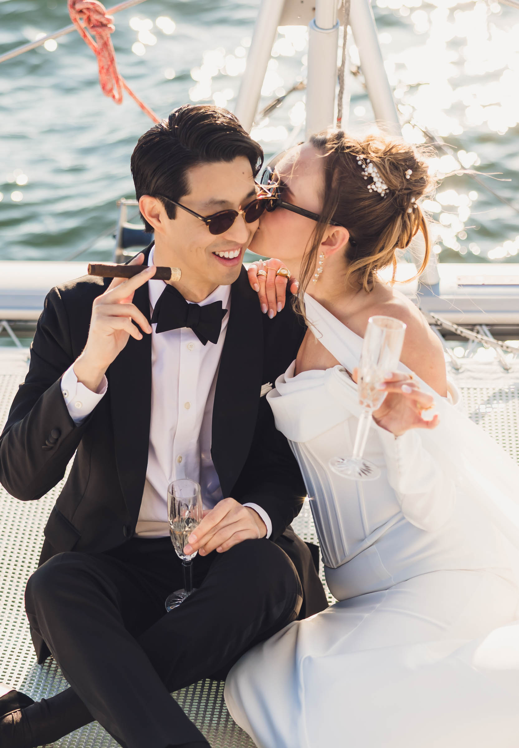 Bride kissing groom on the cheek while holding champagne on a sailboat, groom holding a cigar—joyful waterfront wedding portrait.