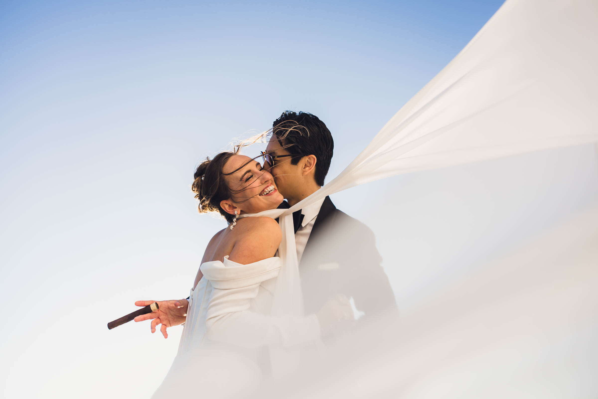 Bride and groom embracing outdoors with veil blowing dramatically in the wind, groom holding a cigar—playful, stylish wedding portrait against a clear sky.