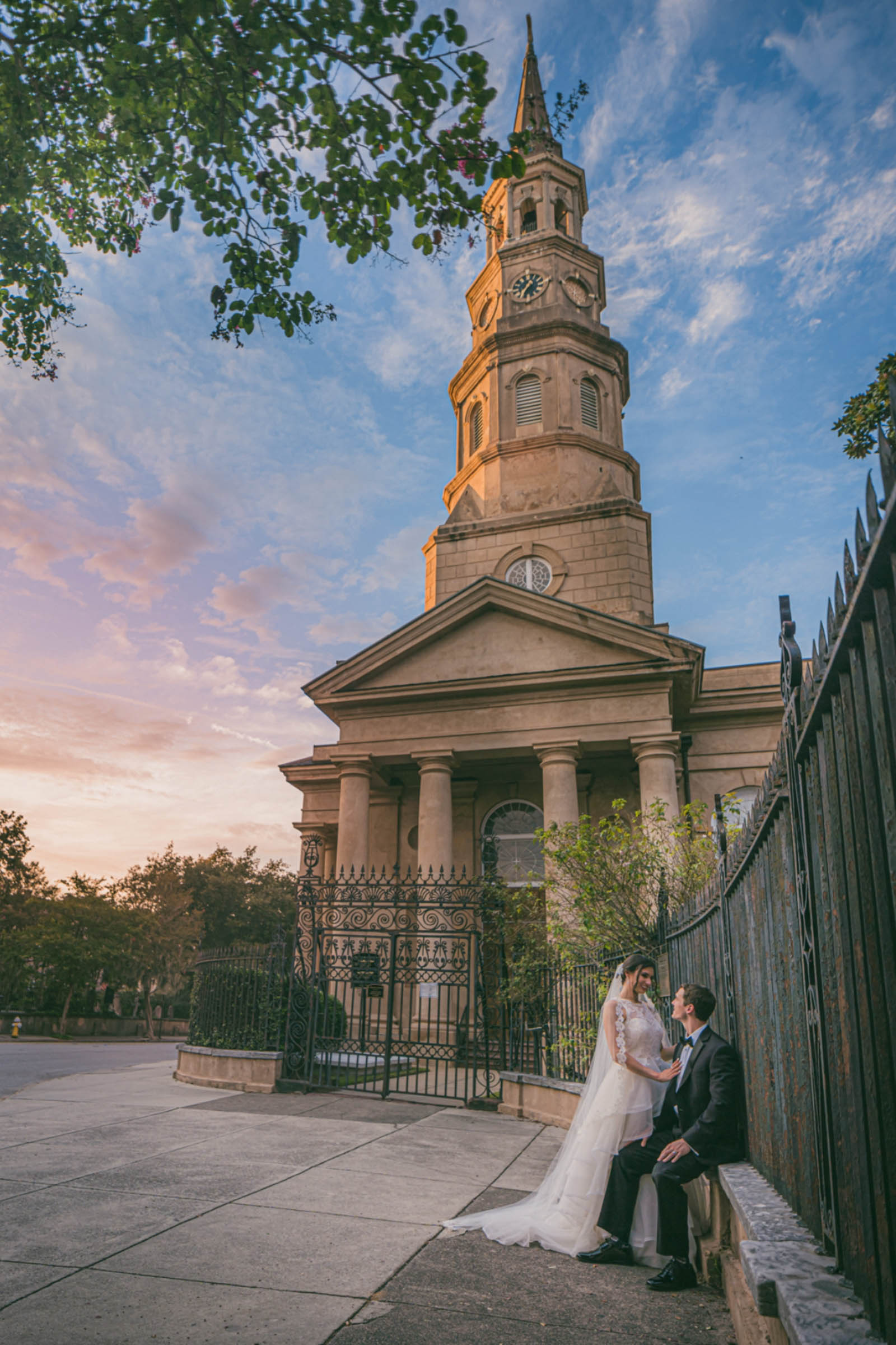 A Couple on the sidewalk of Downtown Charleston with St. Phillips Church towering above. The groom sits by the iron fence as she touches his chest.