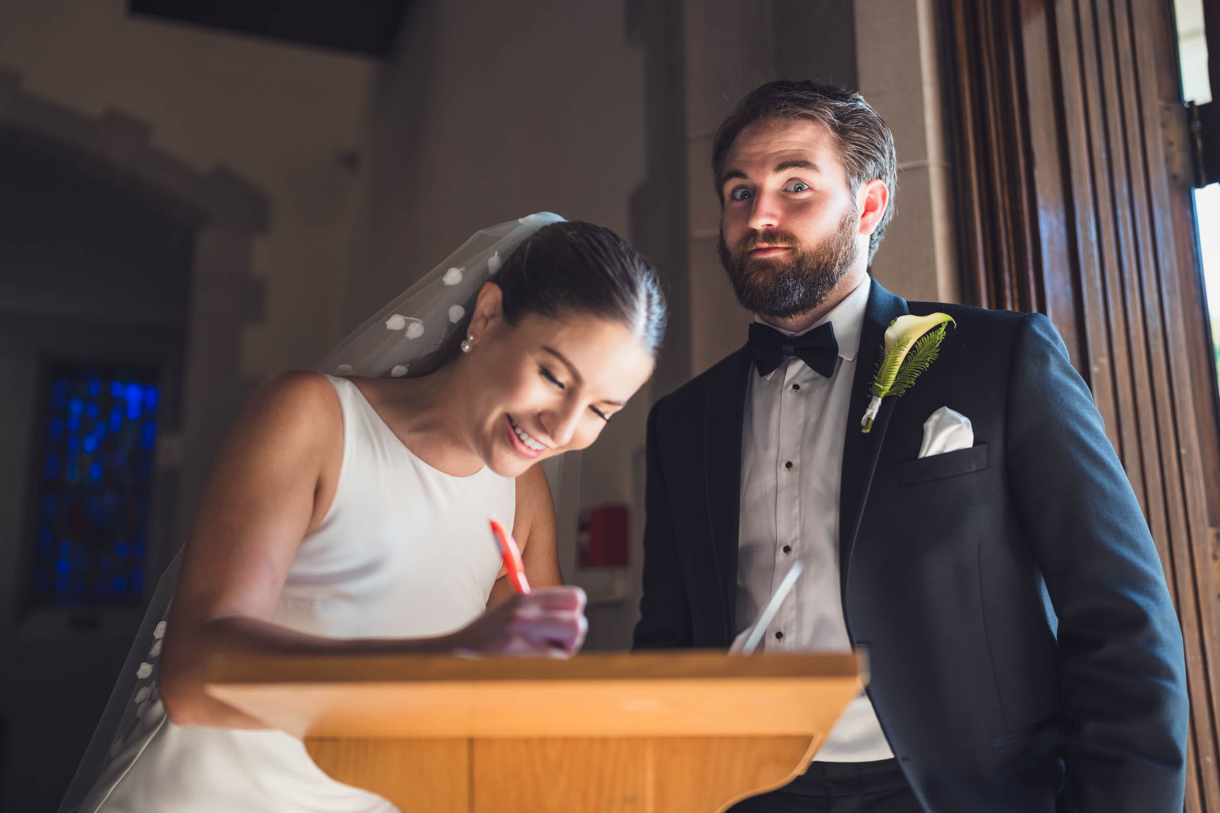 A Photo of a bride and groom signing the Marriage License. The groom looks at the camera with alarm. Standing near the front doorway of Sumerall Chapel at the Citadel.