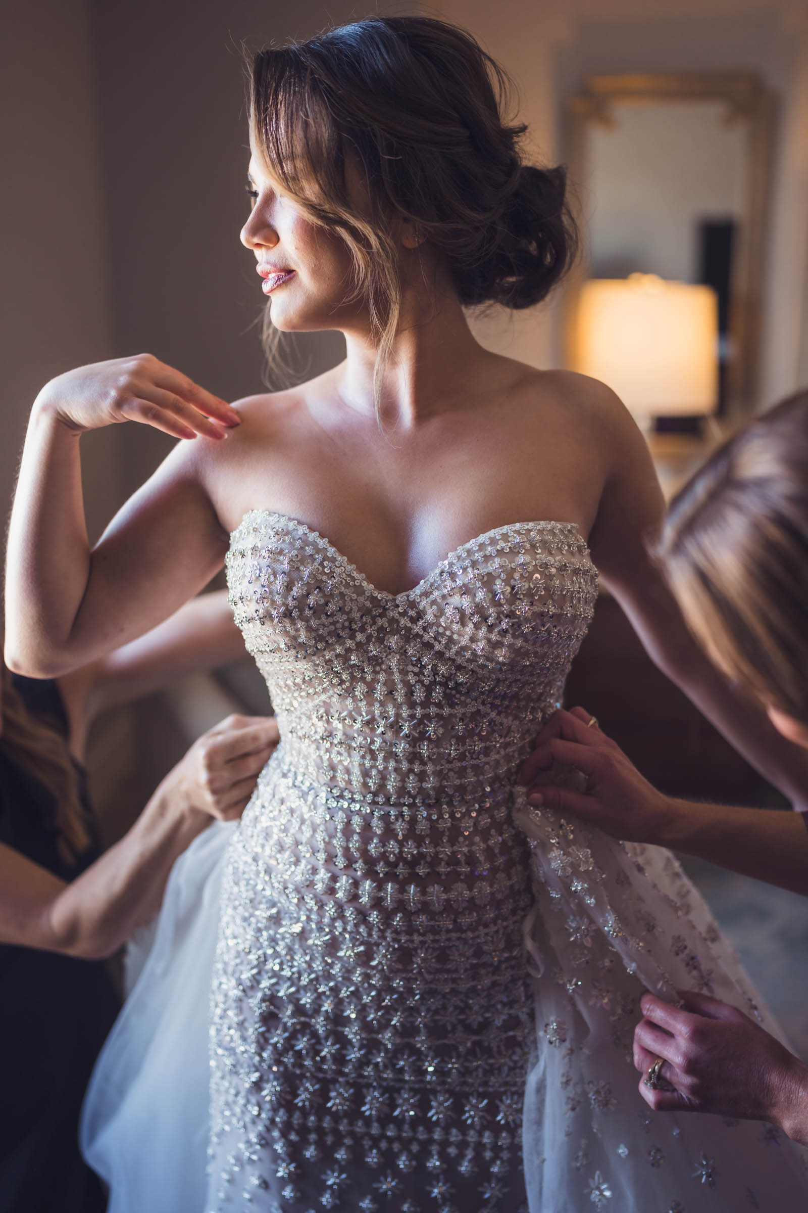 A bride looks towards the light as her ladies attach her train during an elegant getting ready moment.