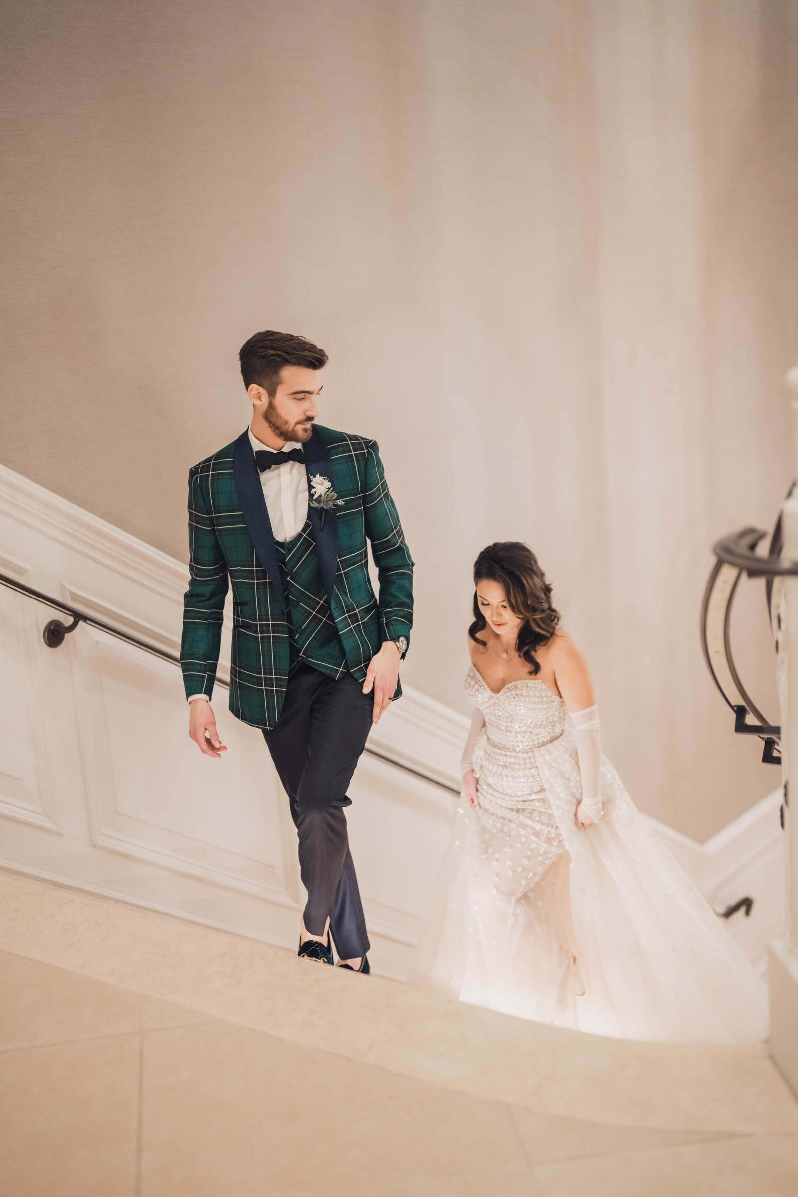 A groom in a green Scottish tartan patterned tuxedo looks back at his bride on the marble stairs at Hotel Bennett