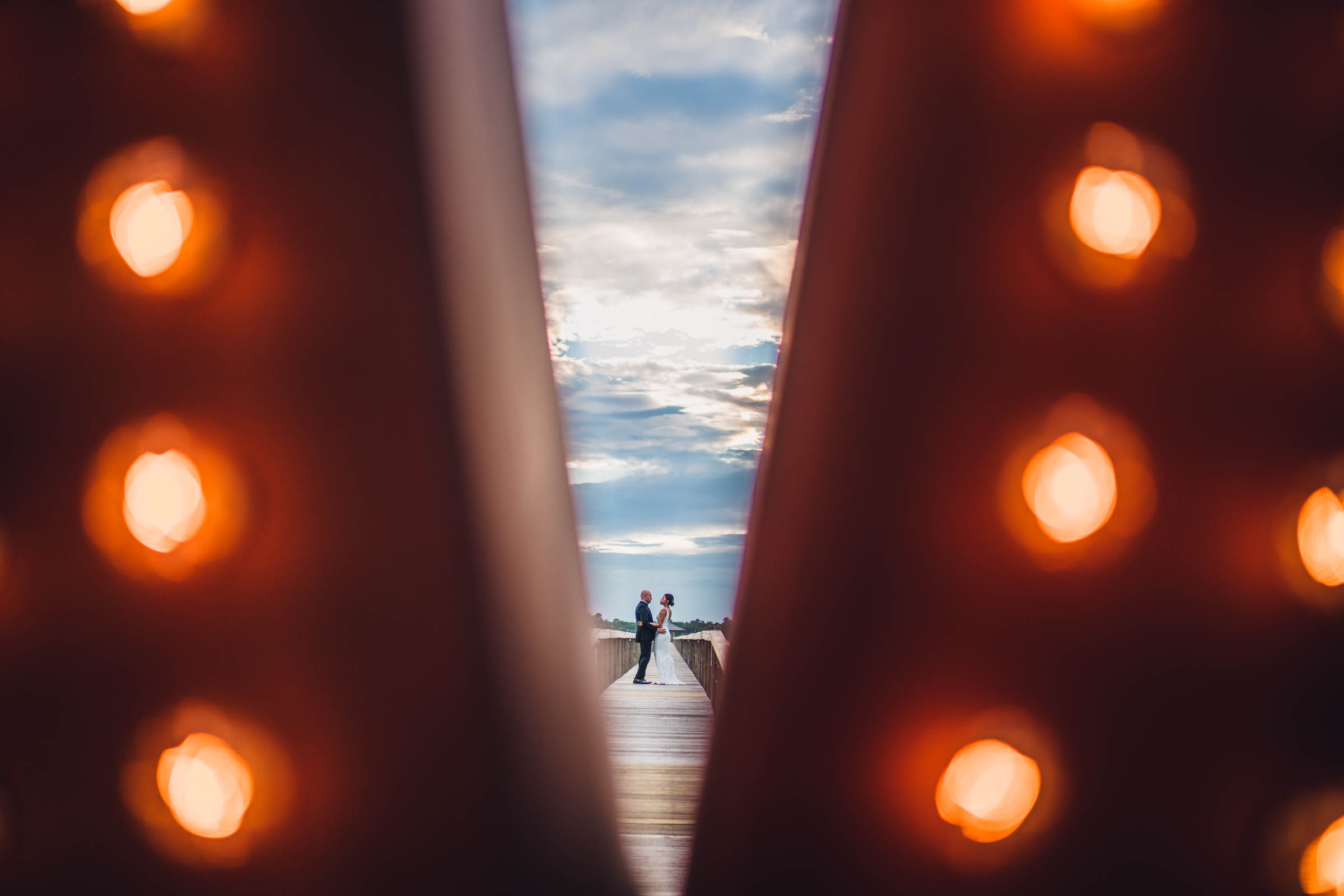 A stylized composition of a couple posing on a dock taken through the letter V of large light marquee letters spelling LOVE.