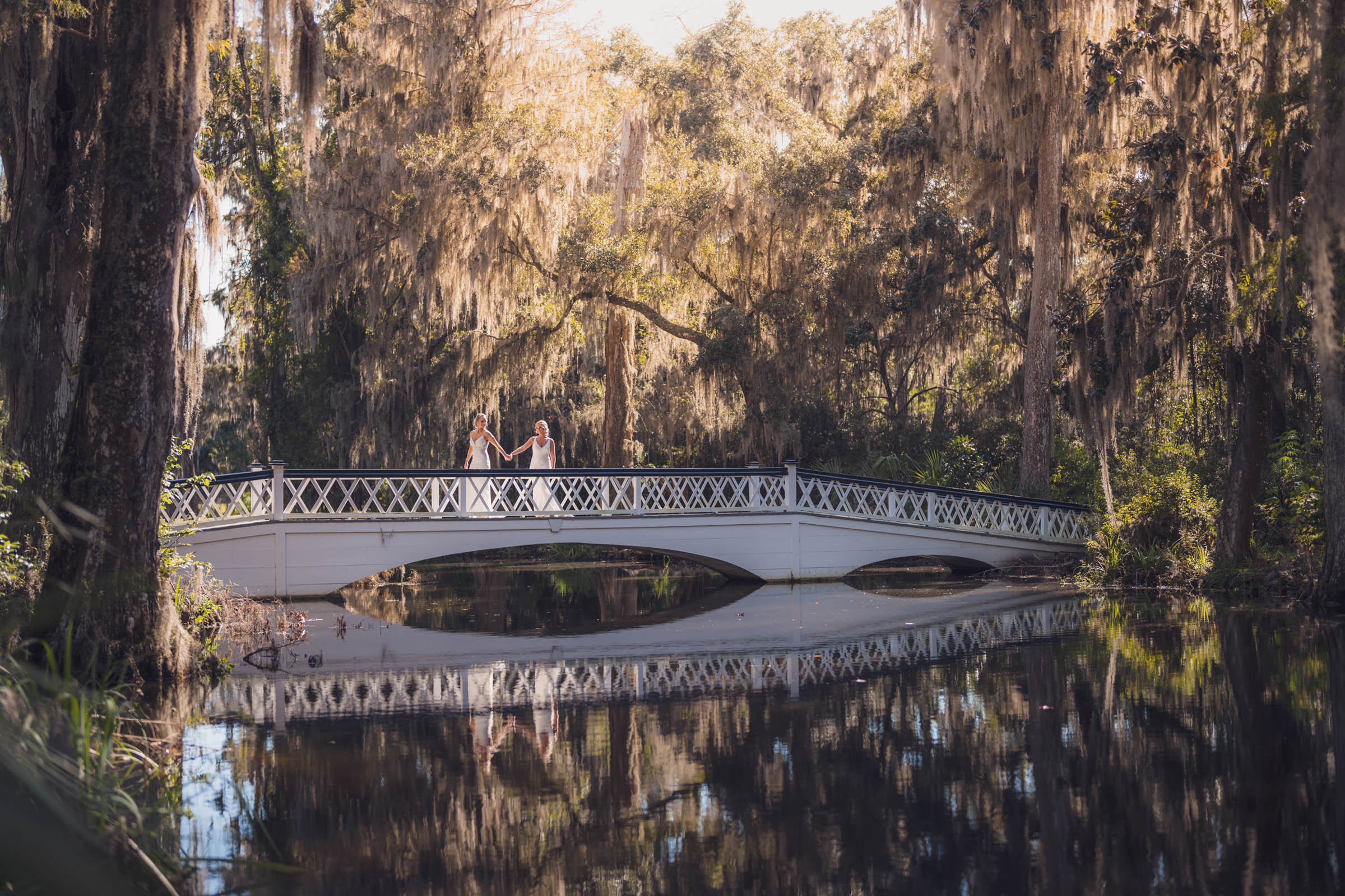 Two bride's hold hands in warm sunlight on the long white bridge at Magnolia Plantation