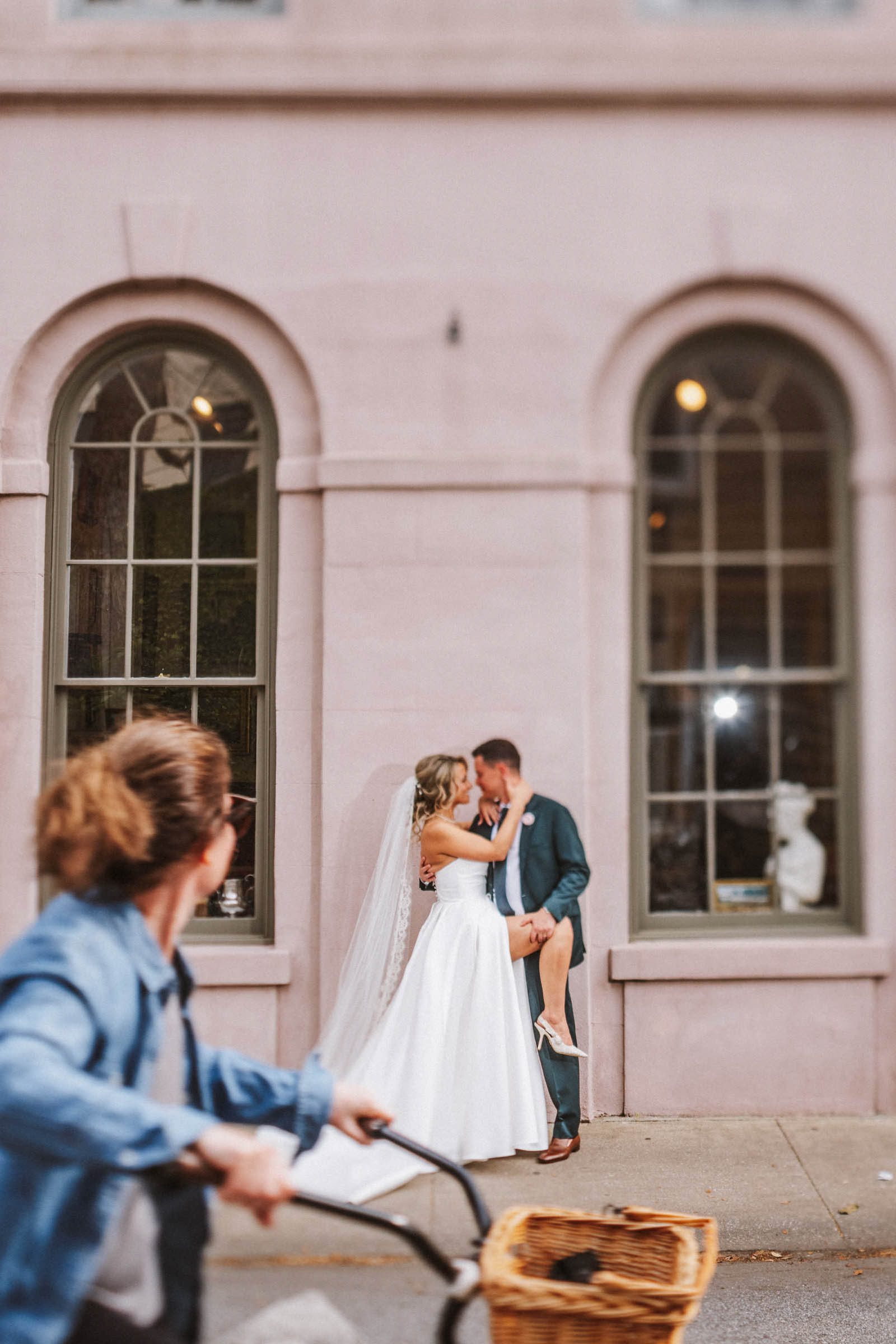 The couple embrace in a dramatic pose, the in front of two arched windows near Mills House Hotel in Charlesotn, SC. A person on a bike is rolling by in the foreground looking at the scene.