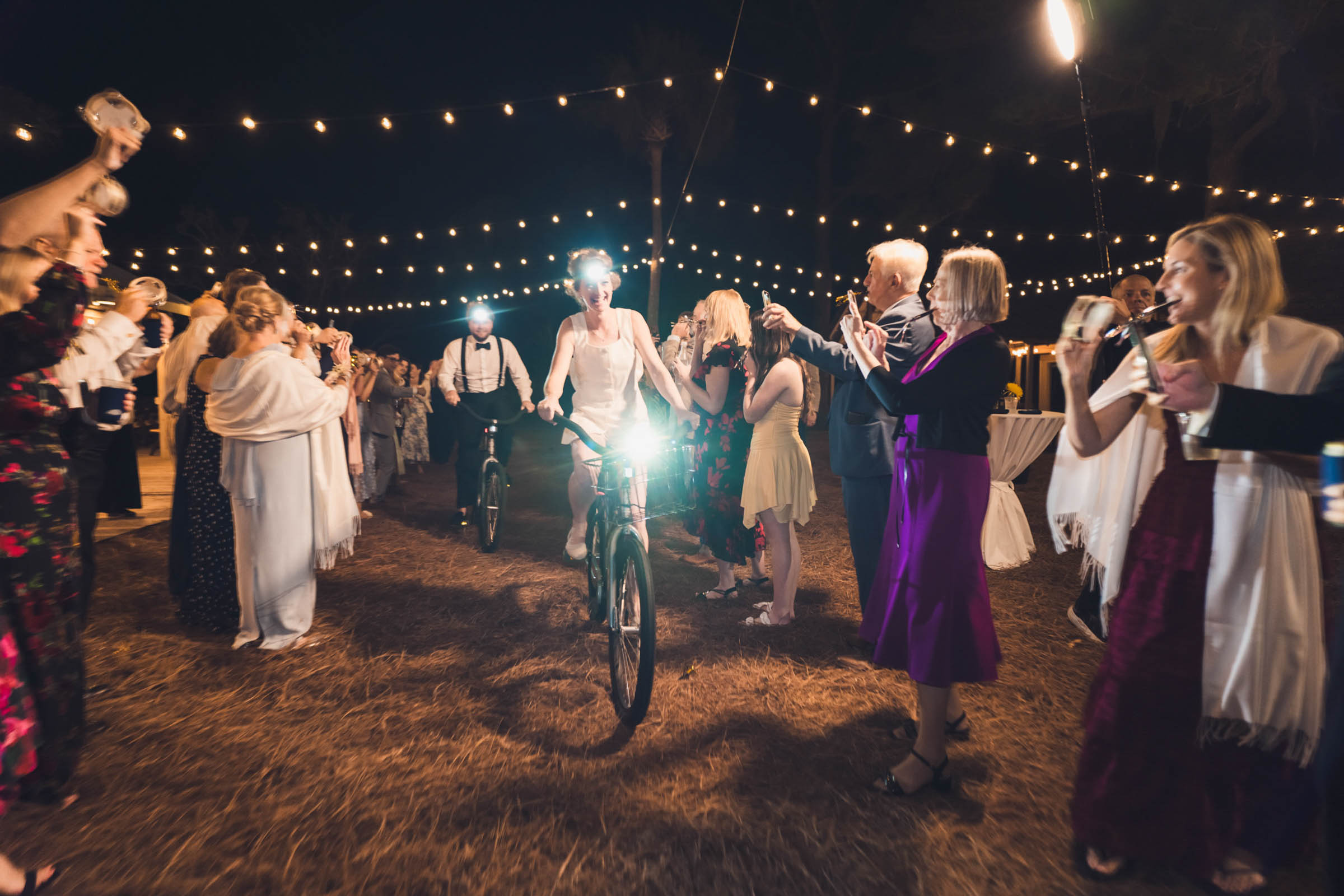 A photo of a bride and groom leaving the reception after dark riding bikes, wearing headlamps as the guests cheer around them