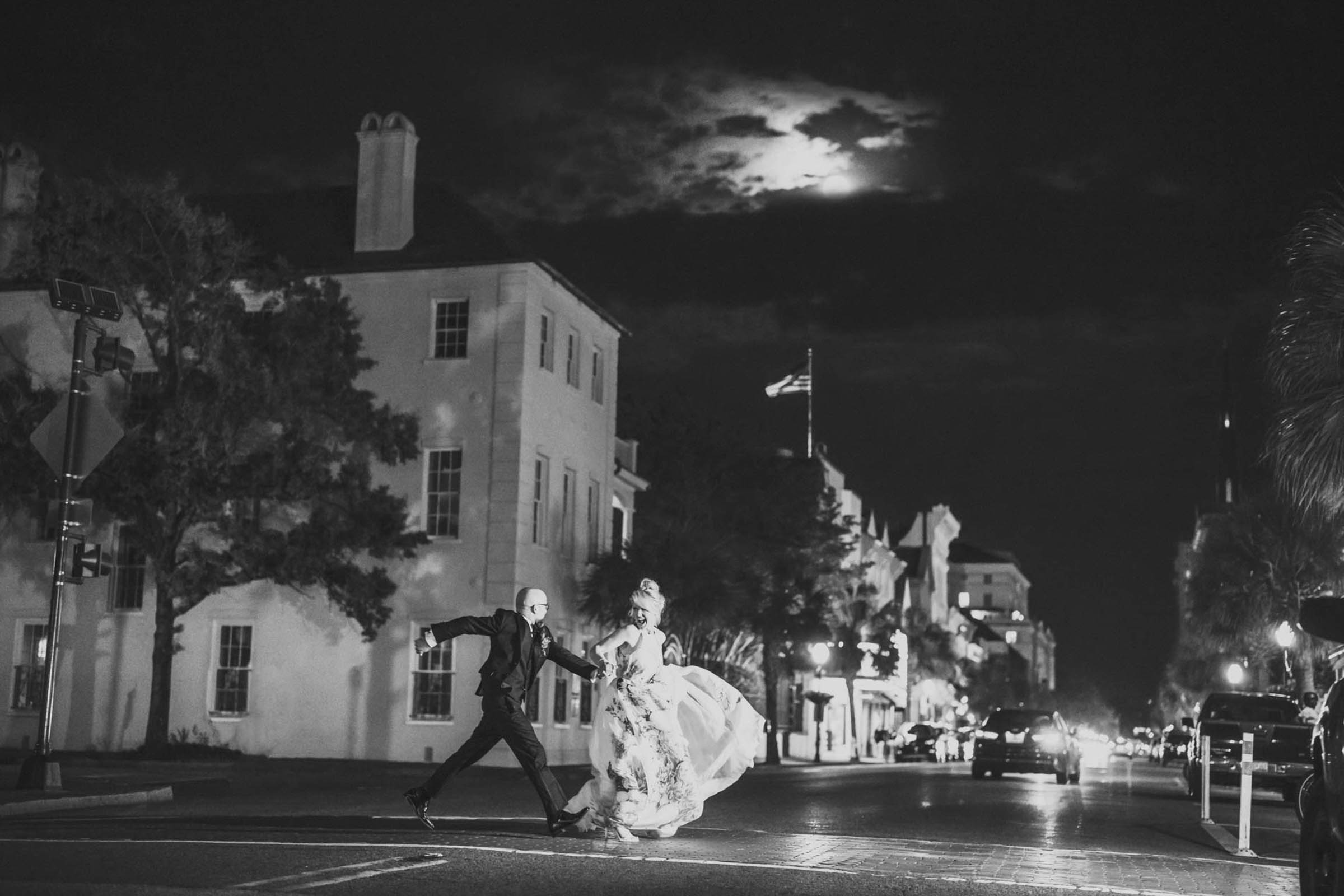 A night photo of a bride and groom crossing King Street in Charleston, SC beneath the moon in a dramatic black and white wedding photograph.