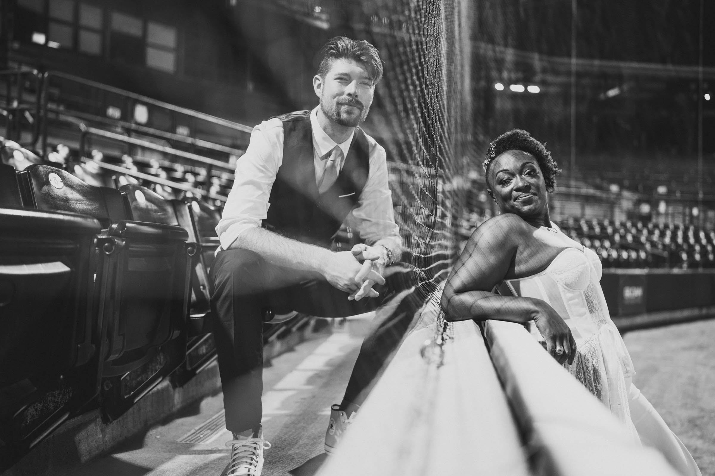 A Black and White photo of a couple posing in Riverdogs Baseball stadium on opposite sides of the backstop net.