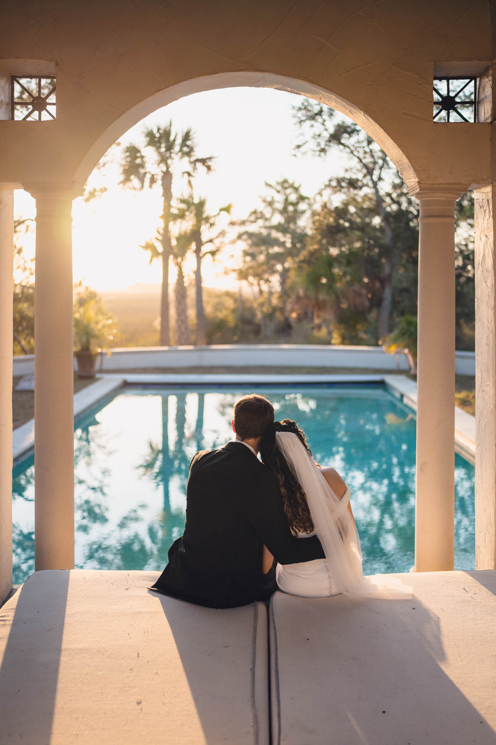 A photo of a bride and groom sitting in a columned cabana, facing away from the camera towards a blue green pool surrounded by palmetto trees