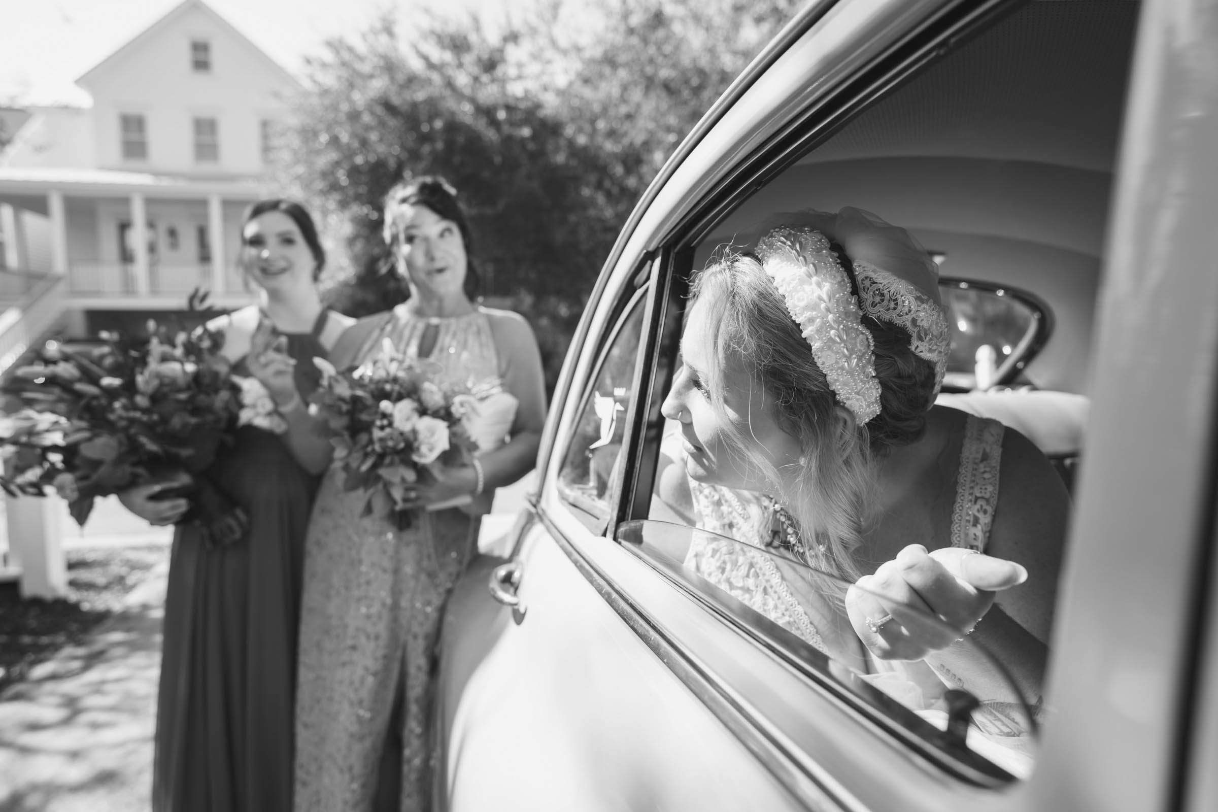 A black and white photo of a bride in a vintage white car looking telling everyone to hurry up. Her mom and sister are visible in the background.
