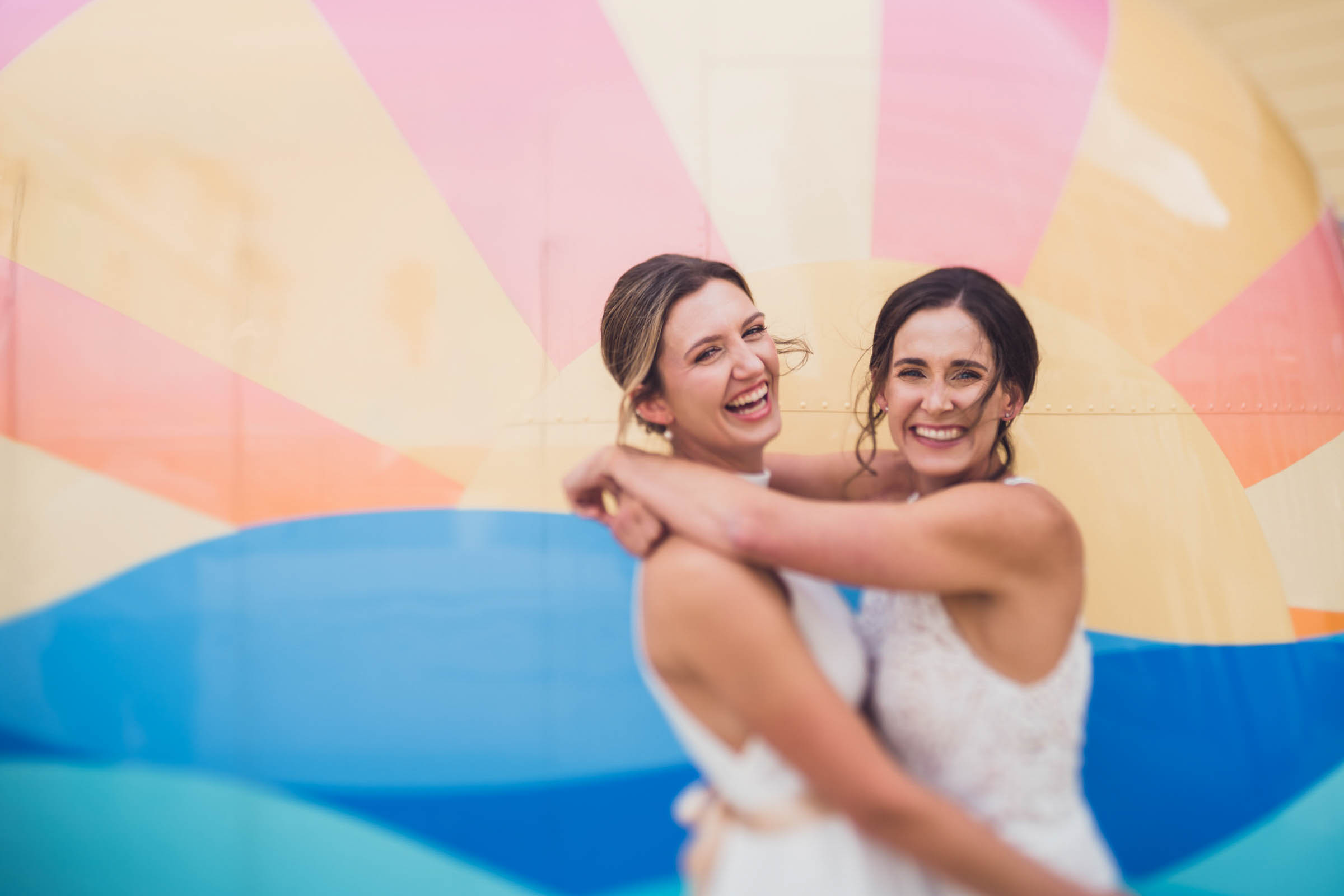 Two brides smile and embrace in front of a colorful mural at Wild Dunes Resort near Charleston, South Carolina.