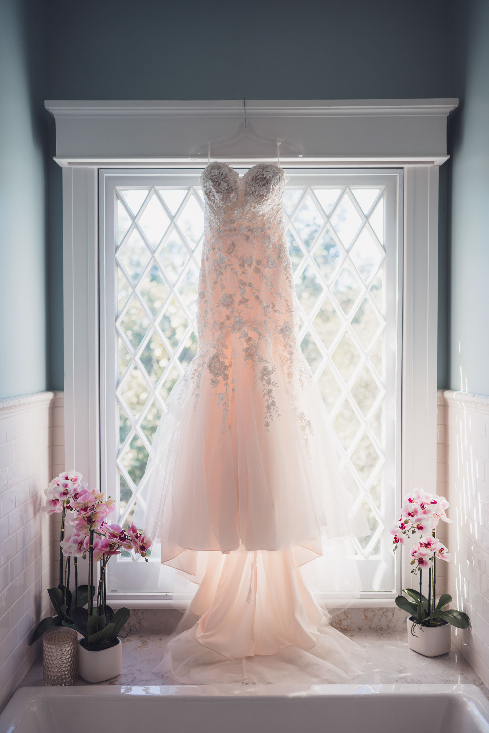 Wedding dress hanging in front of bright window with orchids in bathroom at Admiral’s House