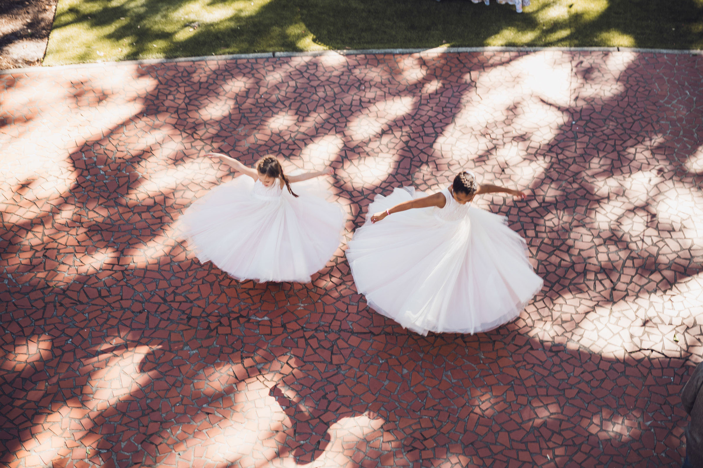 Overhead view of two flower girls spinning in white dresses on brick courtyard with tree shadows