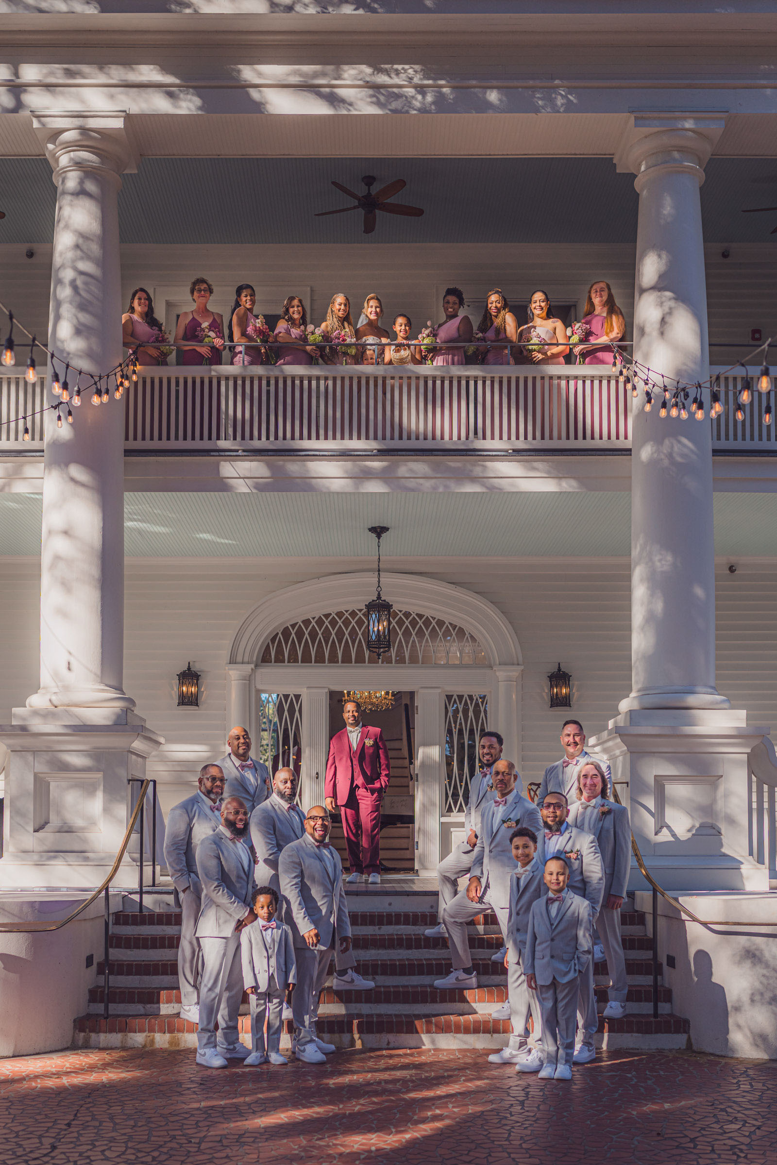 Wedding party group portrait with groom and groomsmen on steps and bridesmaids on balcony of Admiral’s House