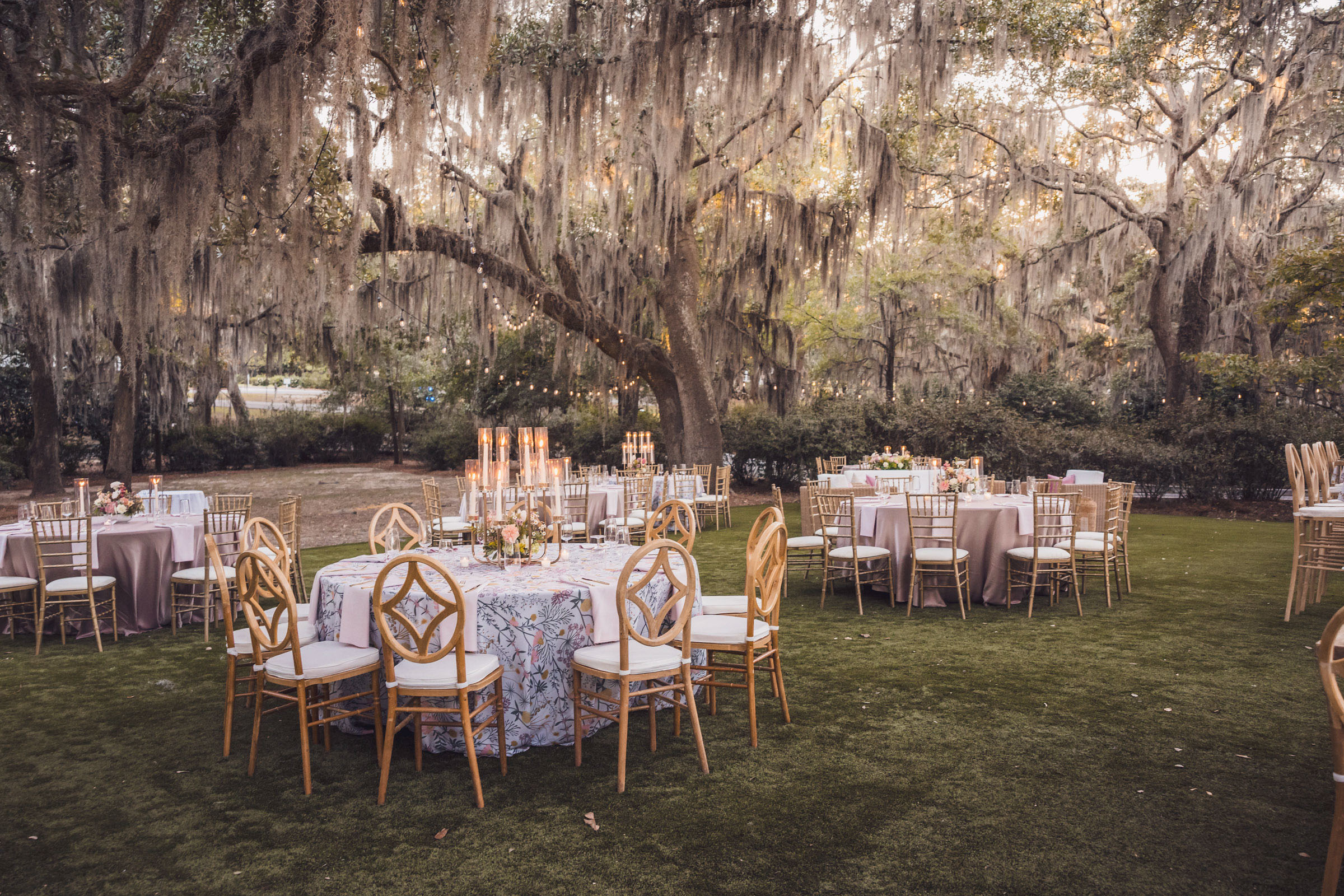 Outdoor wedding reception setup with round tables and string lights beneath oak trees at Admiral’s House