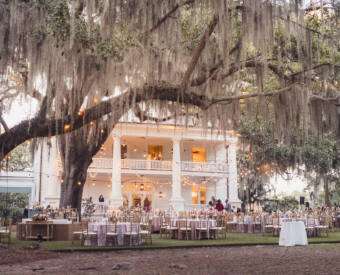 Wide view of outdoor wedding reception at Admiral’s House with string lights and oak trees