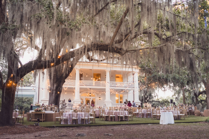 Wide view of outdoor wedding reception at Admiral’s House with string lights and oak trees