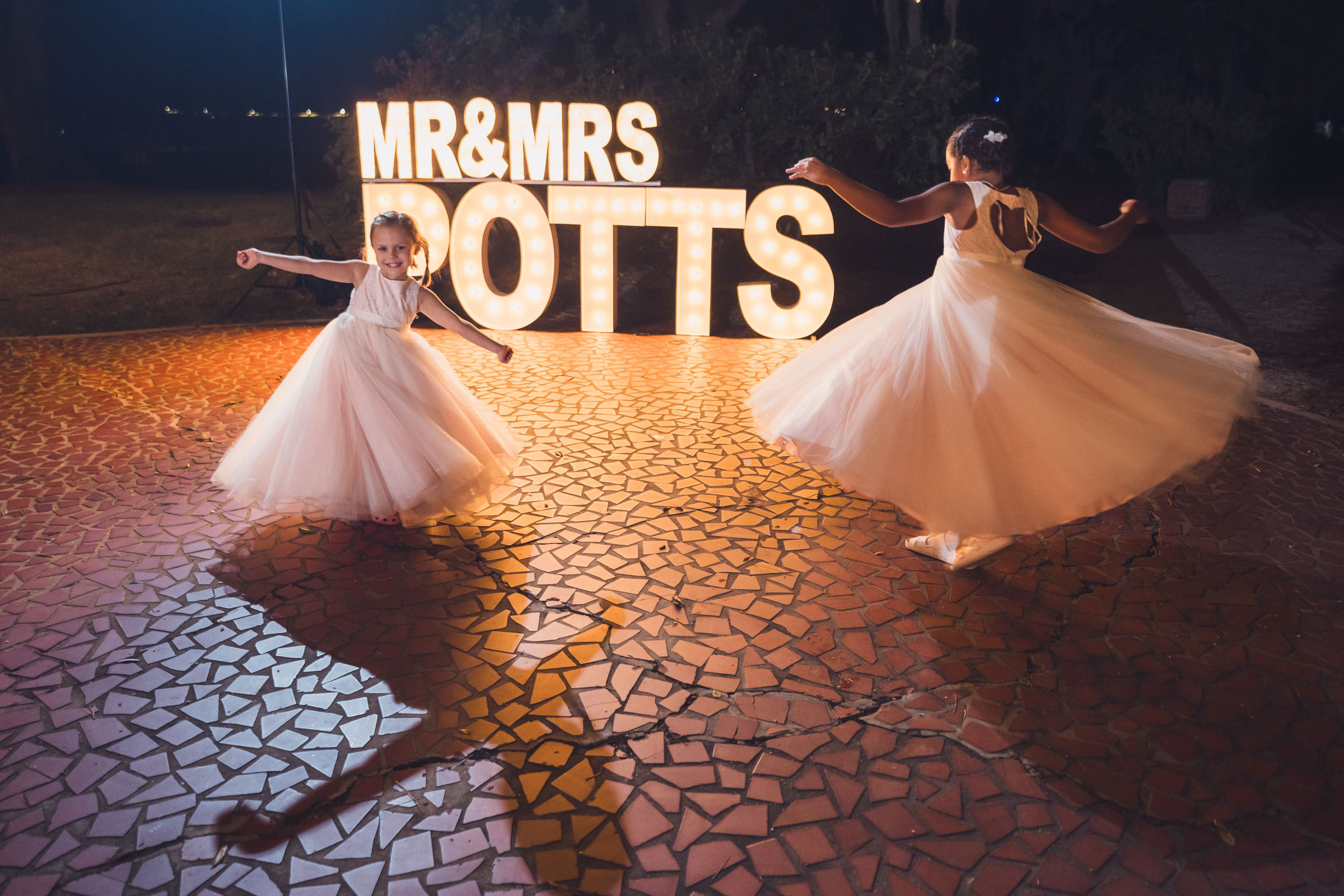 Two flower girls spinning in white dresses in front of illuminated “Mr & Mrs Potts” sign at outdoor wedding reception