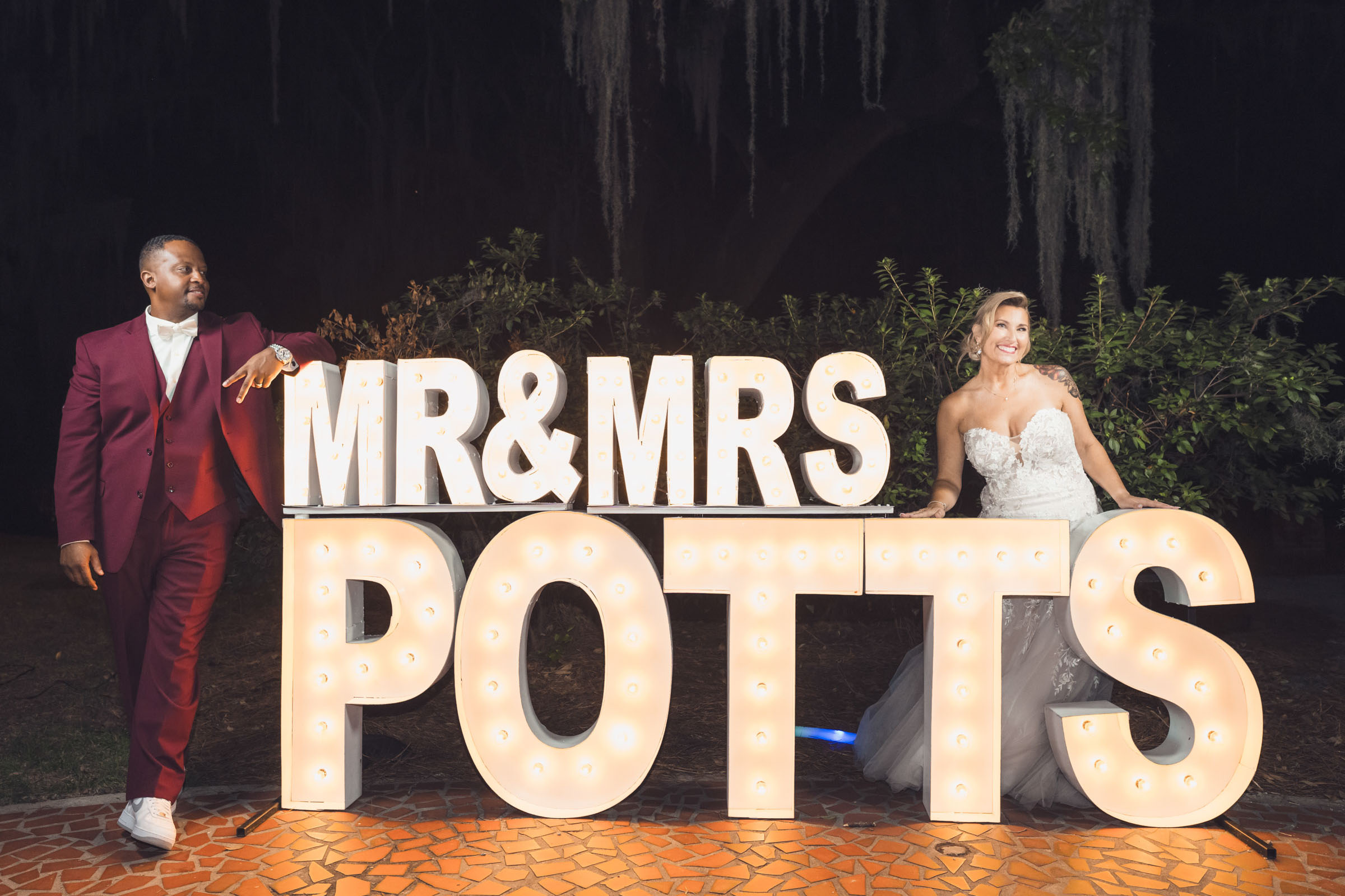 Bride and groom posing with illuminated “Mr & Mrs Potts” sign at nighttime wedding reception