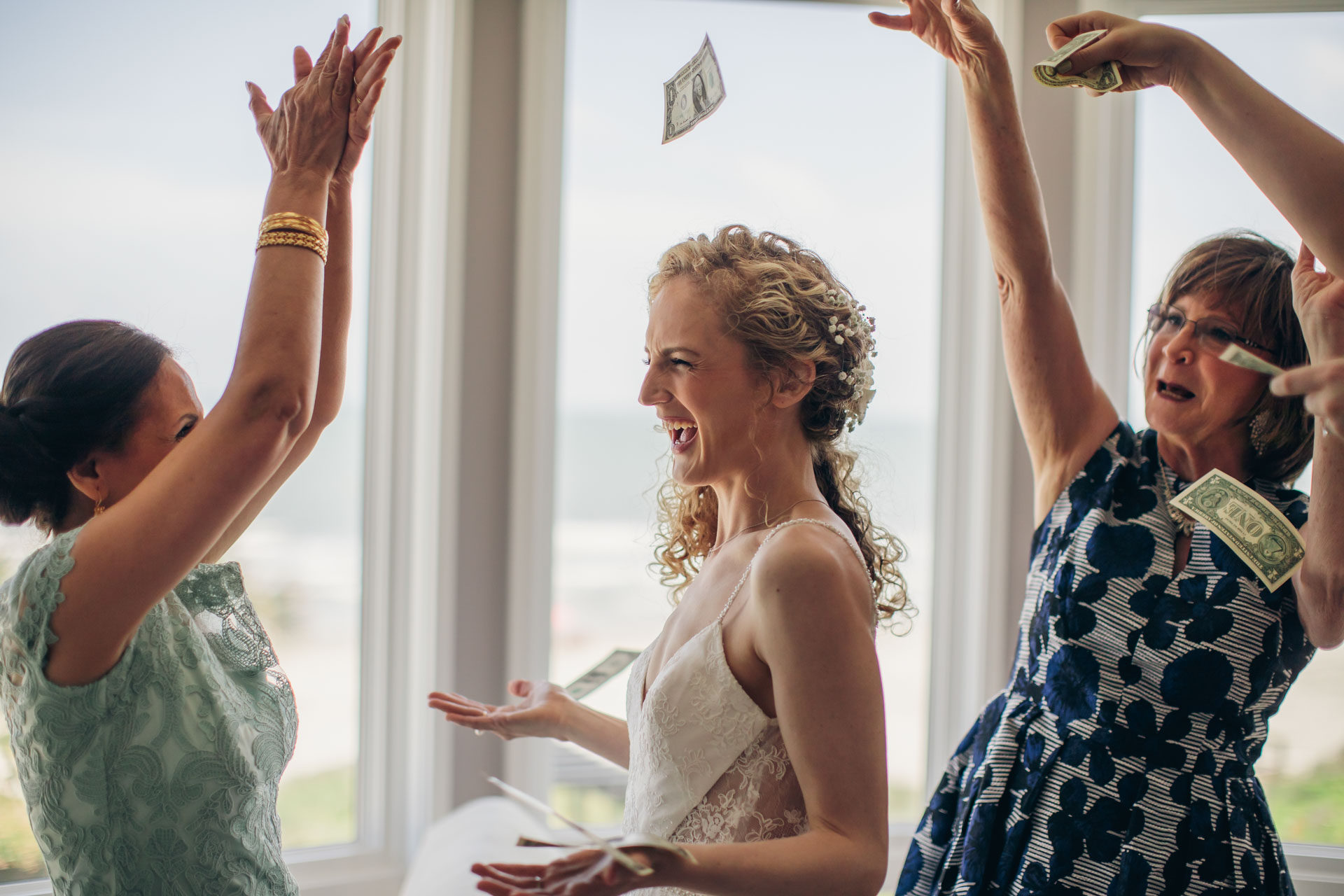 Bride laughing as women around her toss dollar bills in the air during a lively getting-ready moment by a window.