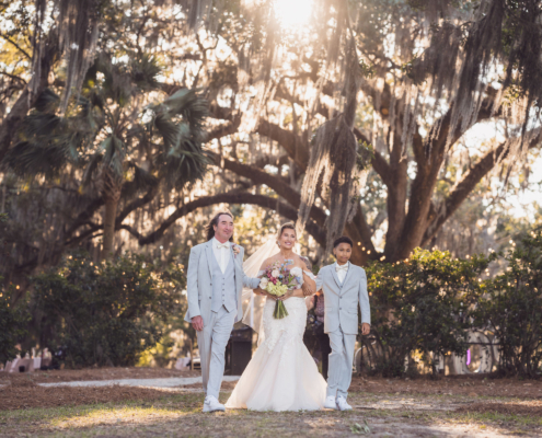 Bride walking with family beneath Spanish moss-covered live oaks on the way to her outdoor wedding ceremony.