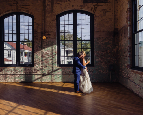 Bride and groom embracing in late-afternoon sunlight beneath the tall factory windows and exposed brick of The Cedar Room.