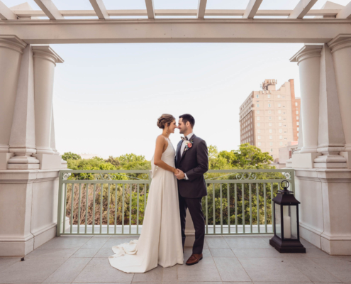 Bride and groom stand beneath the columns of a Hotel Bennett piazza overlooking downtown Charleston.
