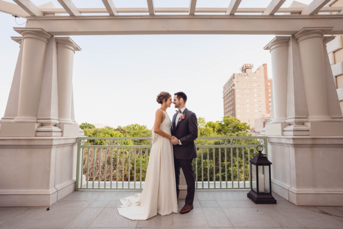 Bride and groom stand beneath the columns of a Hotel Bennett piazza overlooking downtown Charleston.